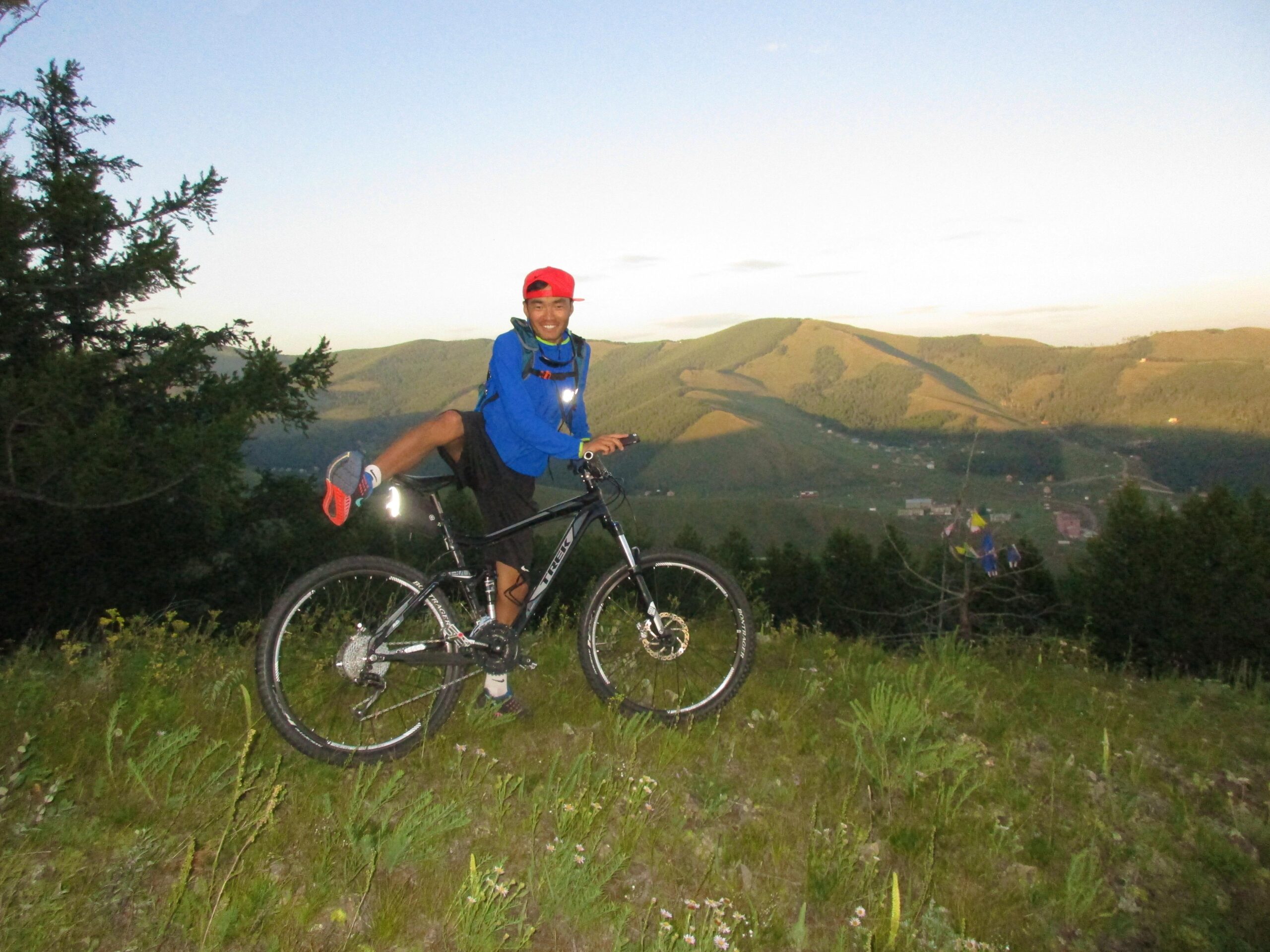 A smiling person wearing a red cap and blue jacket poses on a mountain bike with one leg raised, set against a scenic backdrop of rolling green hills and a clear sky. The setting suggests a late afternoon or early evening scene, with grass and trees surrounding the cyclist. Selkh Am mountain bike trail.