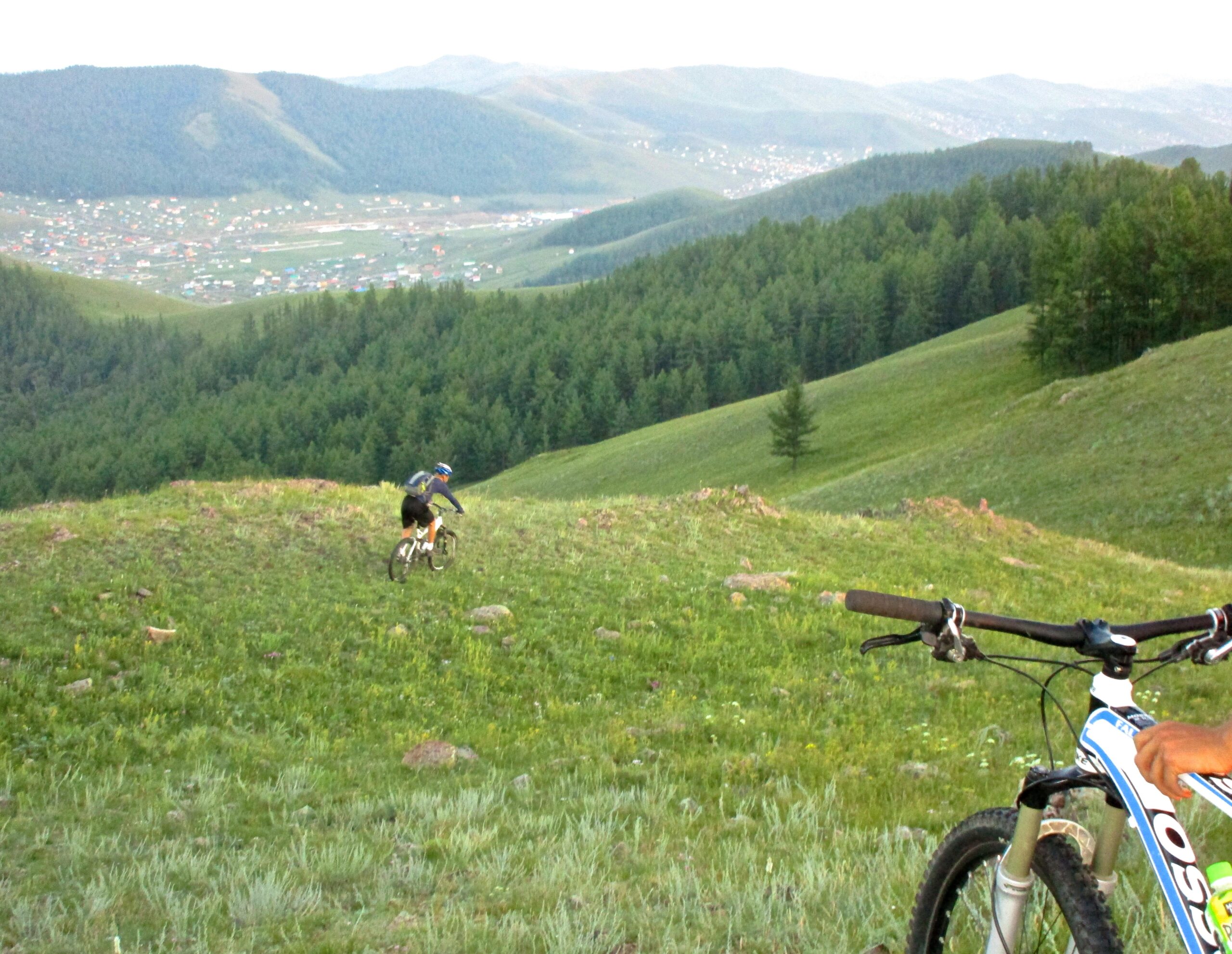 A mountain biker descends a grassy hill with rocky terrain, surrounded by lush green trees and rolling mountains in the background. A second bike is positioned in the foreground, emphasizing the adventure and natural beauty of the landscape. Selkh Am mountain bike trail.