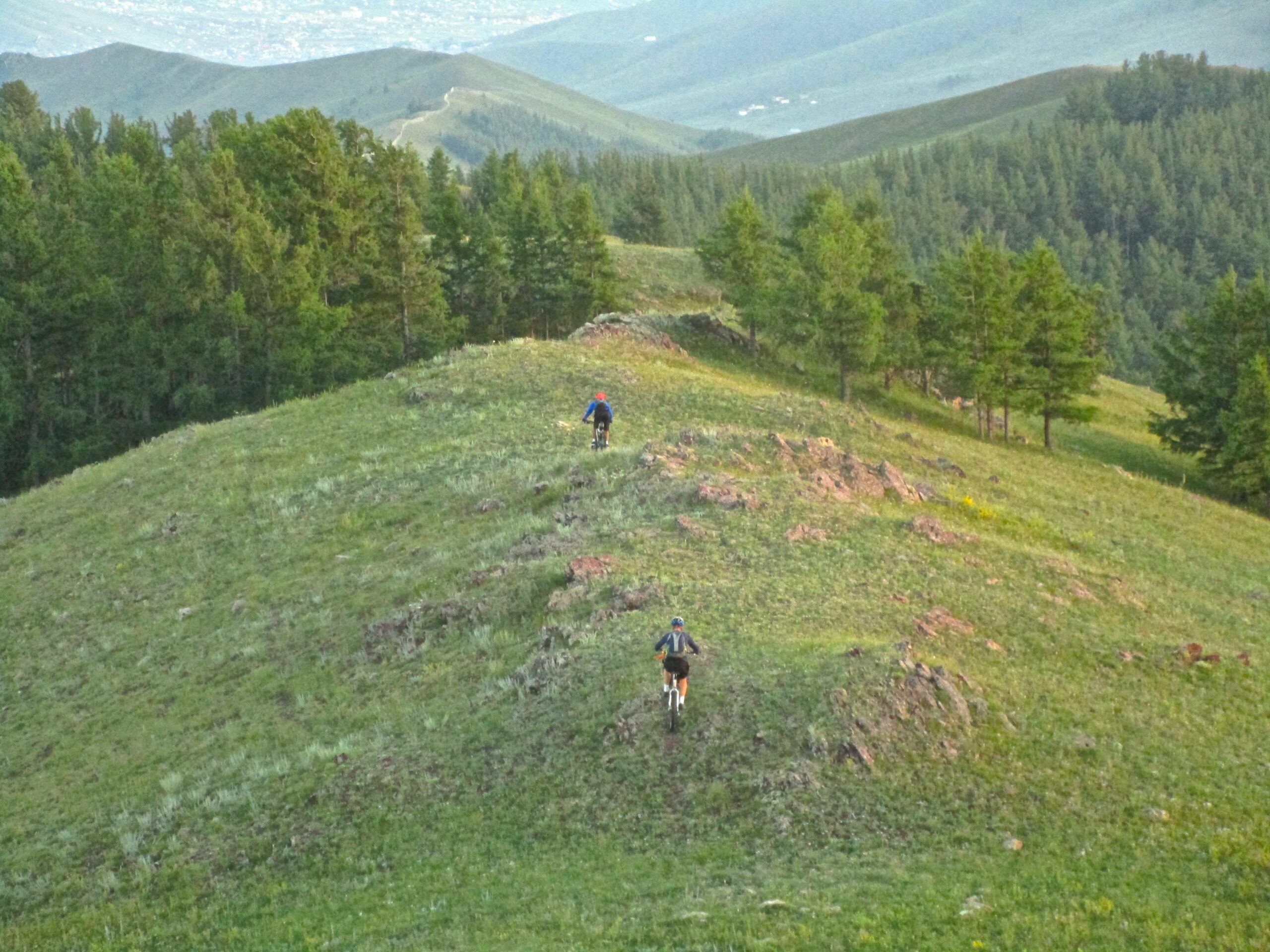 Two mountain bikers riding on a grassy hillside, surrounded by trees, with a view of distant mountains and a valley below. The scene captures a serene outdoor environment, ideal for cycling and exploring nature. Selkh Am mountain bike trail.
