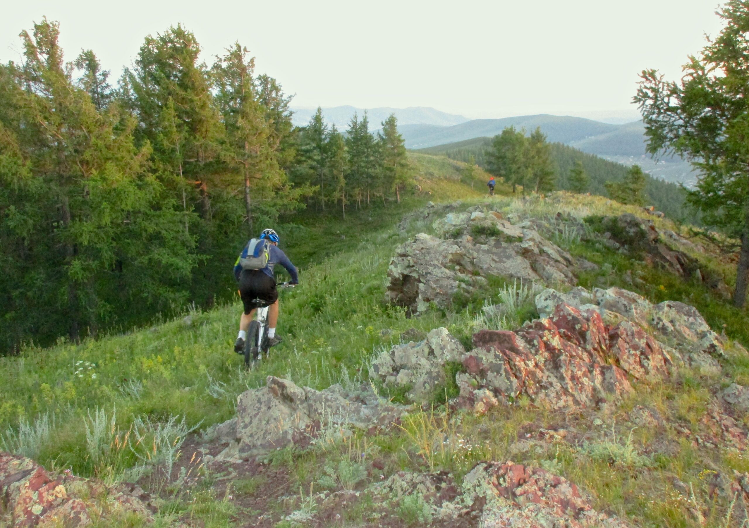 A mountain biker navigates a grassy trail surrounded by rocks and trees, with rolling hills and mountains visible in the background. Another cyclist is seen in the distance, enjoying the scenic outdoor landscape. The scene captures the essence of outdoor adventure and nature exploration. Selkh Am mountain bike trail.