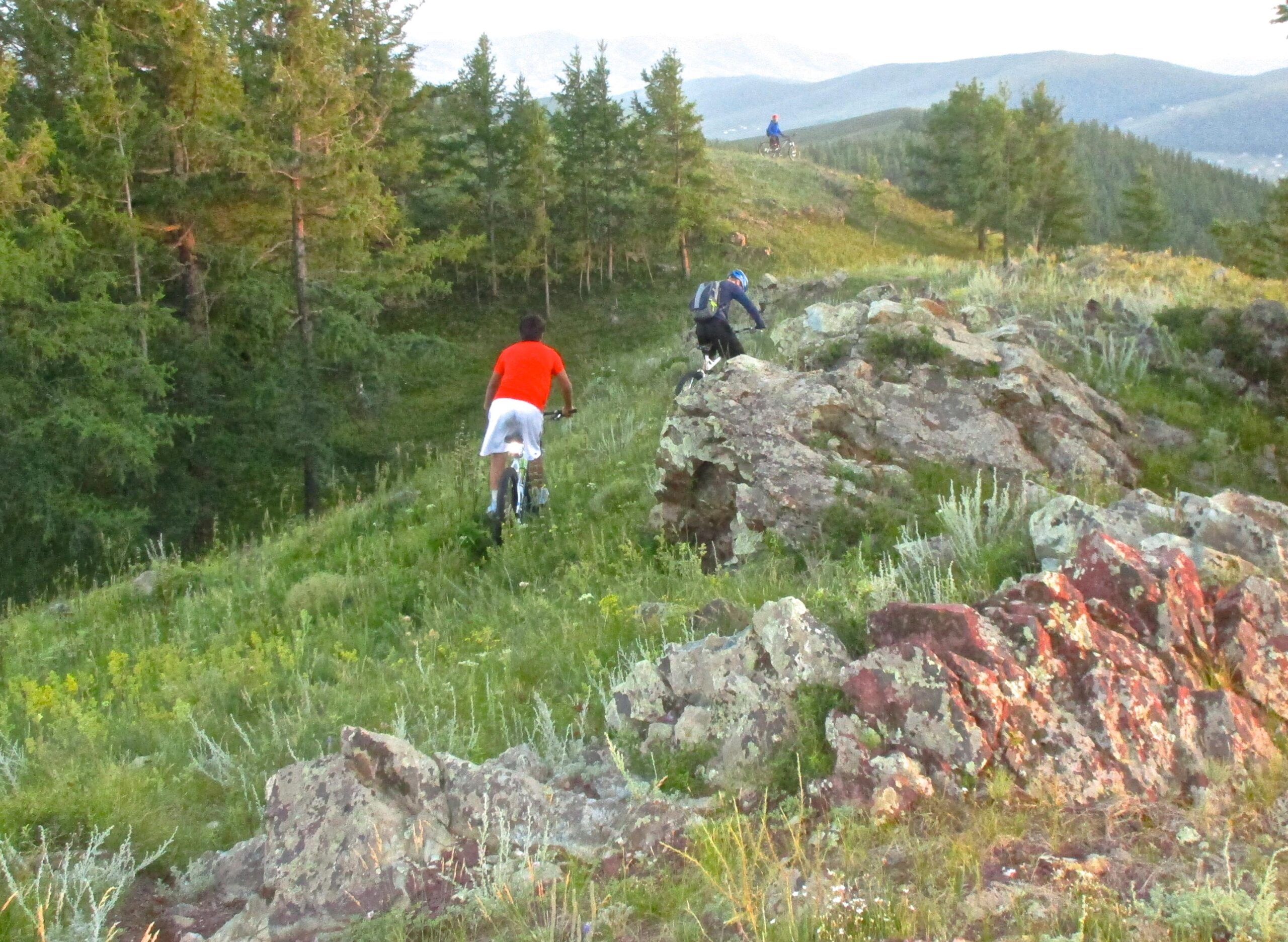 Two mountain bikers riding on a grassy trail surrounded by trees and rocky terrain. One biker, wearing an orange shirt and white shorts, is in the foreground, while another biker in blue is further up the hill. The scene is set in a natural environment with distant mountains visible in the background. Selkh Am mountain bike trail.