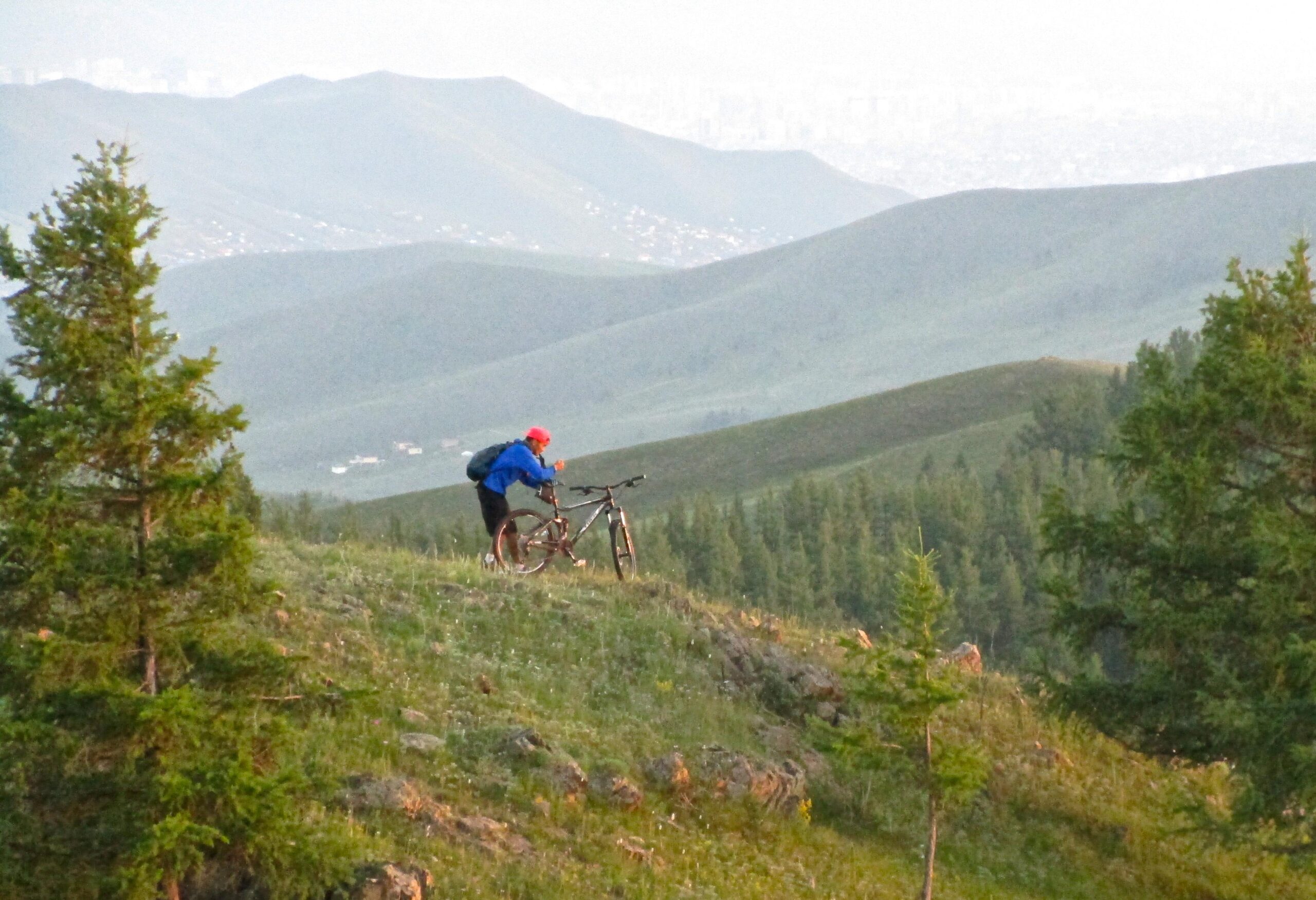 A cyclist in a blue jacket and red helmet stands beside a mountain bike on a grassy hillside, surrounded by rolling green mountains and trees. The background features a distant city skyline, partially obscured by mist. Selkh Am mountain bike trail.