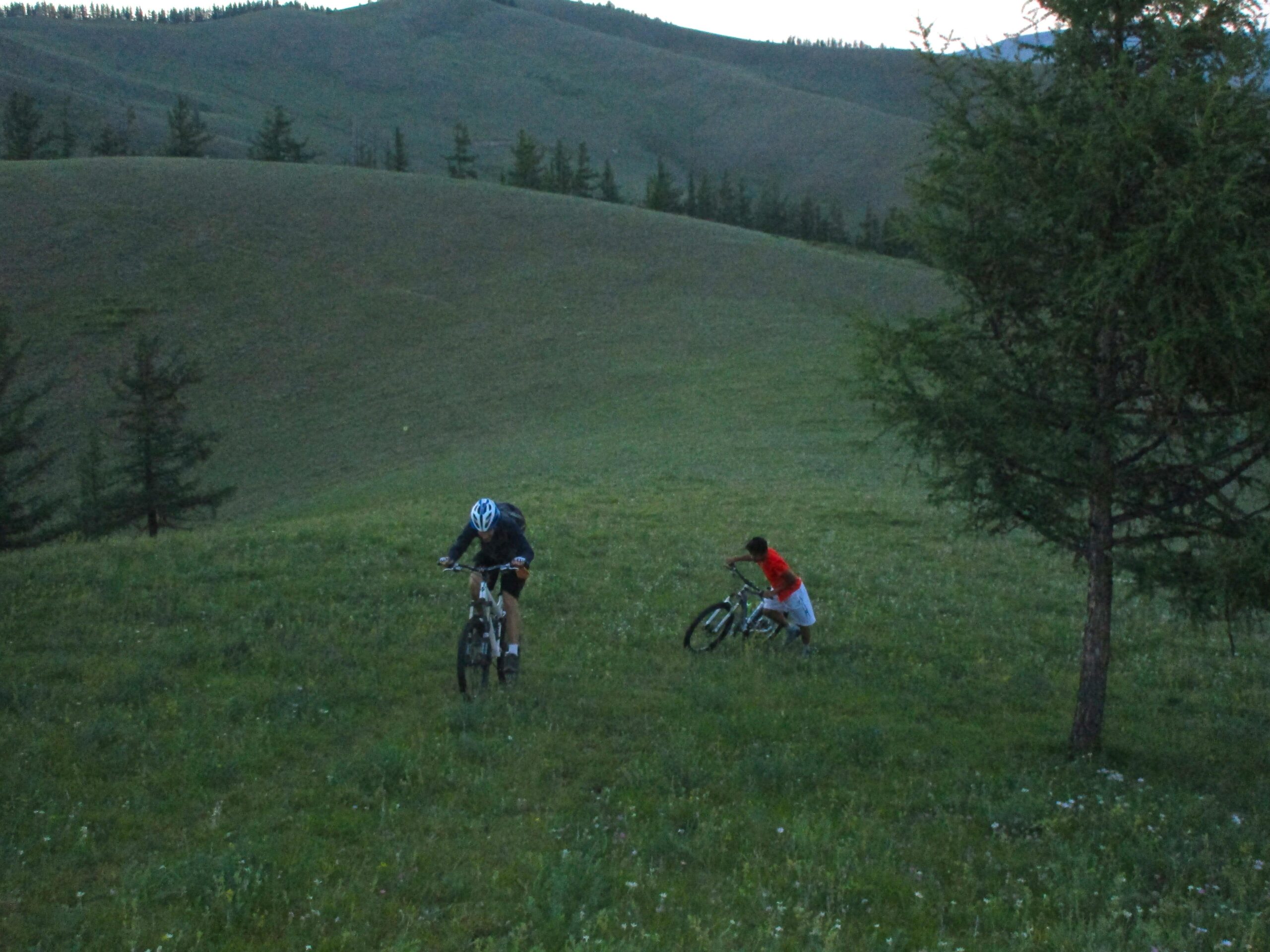 Two cyclists navigating a grassy hill in a rural landscape, with rolling green hills in the background and scattered trees. One cyclist is in the foreground, wearing a helmet and focused on managing their bike, while the other cyclist is further back, dressed in a red shirt, adjusting their bicycle. The scene captures the beauty of outdoor cycling in nature during the evening. Selkh Am mountain bike trail.