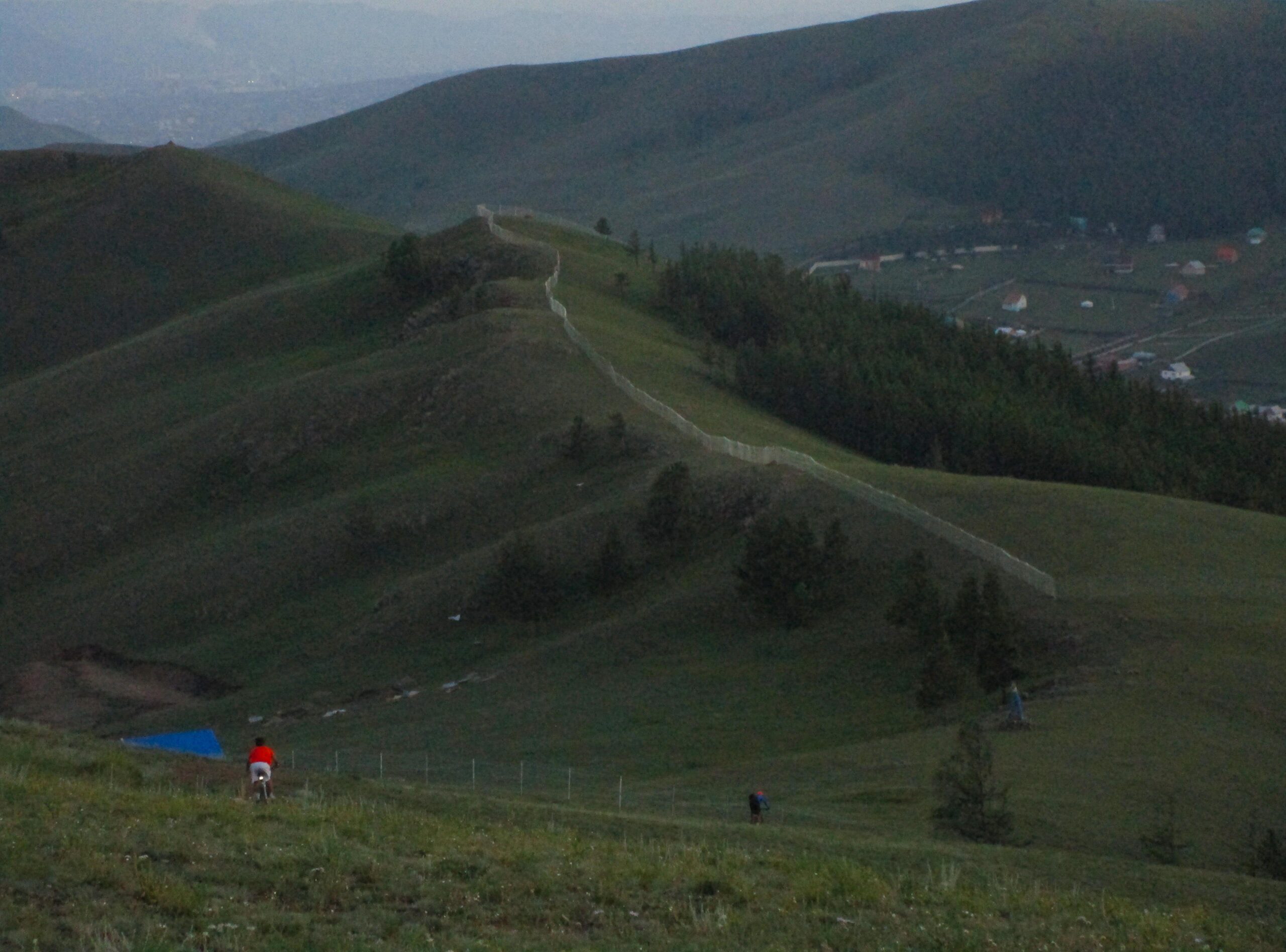 A scenic view of rolling green hills under a twilight sky, featuring a winding fence that traverses the landscape. In the foreground, two individuals are seen walking, one wearing a red shirt near the bottom left, while the other is further up the hill. A blue tarp is located on the ground, surrounded by grass and sparse trees, with a small village visible in the distance. Selkh Am mountain bike trail.