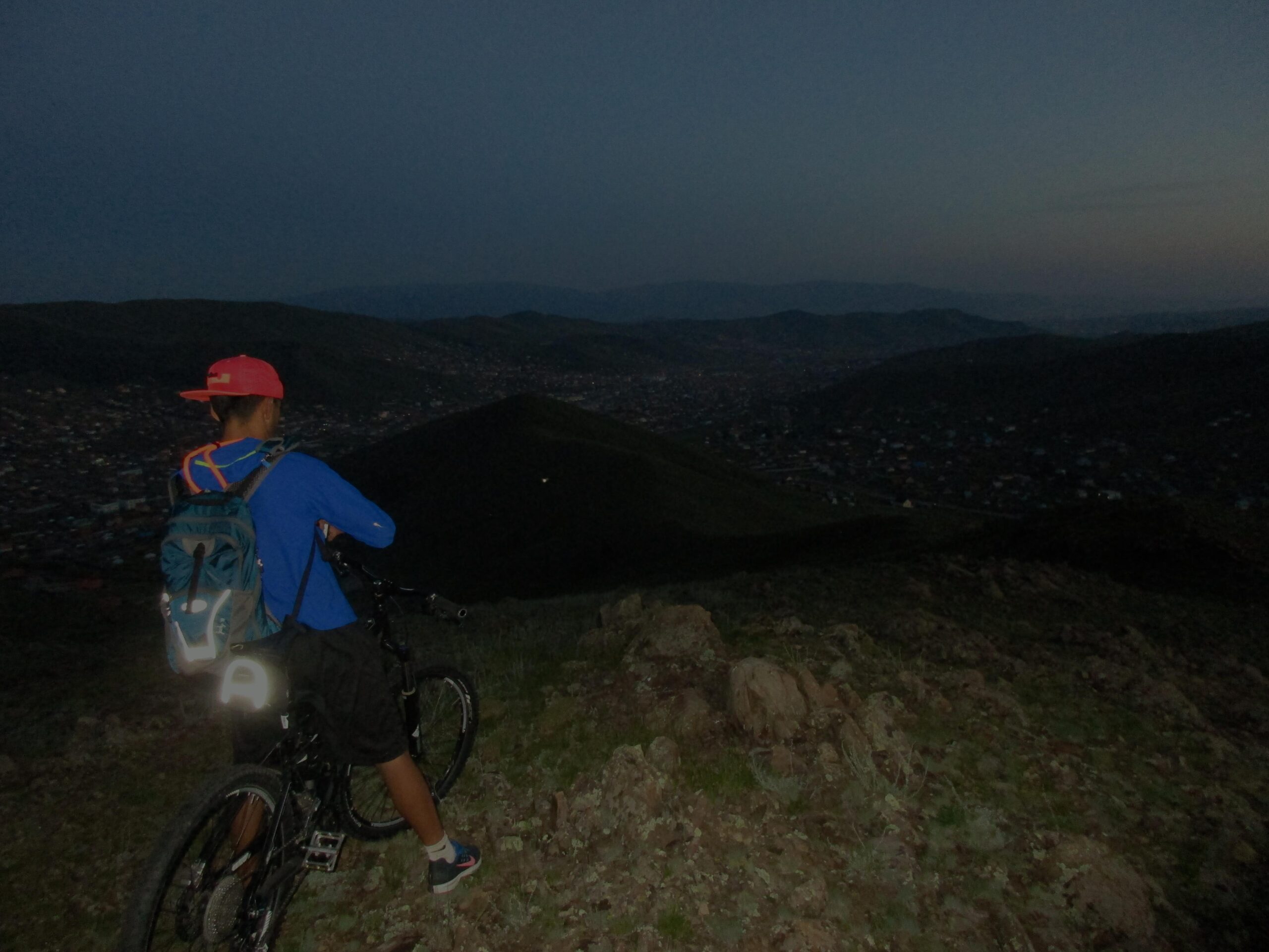 A person wearing a blue long-sleeve shirt and a red cap is standing on a hillside with a mountain bike, overlooking a valley at dusk. The landscape is dimly lit, with distant lights from a town visible below. The individual is equipped with a backpack, and the bike has a reflective light. Selkh Am mountain bike trail.