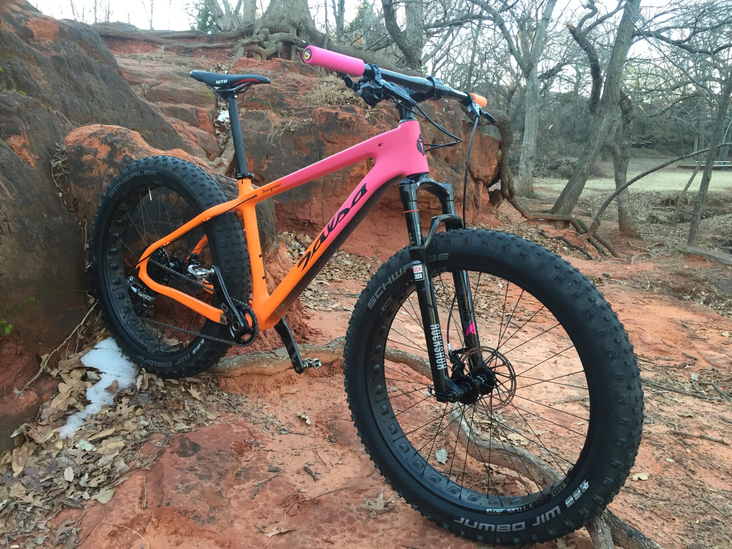 Salsa Beargrease: A colorful fat bike with a pink and orange frame rests against a rocky surface, surrounded by trees and a patch of grass. The bike features thick tires designed for rugged terrain, showcasing a blend of nature and outdoor adventure.