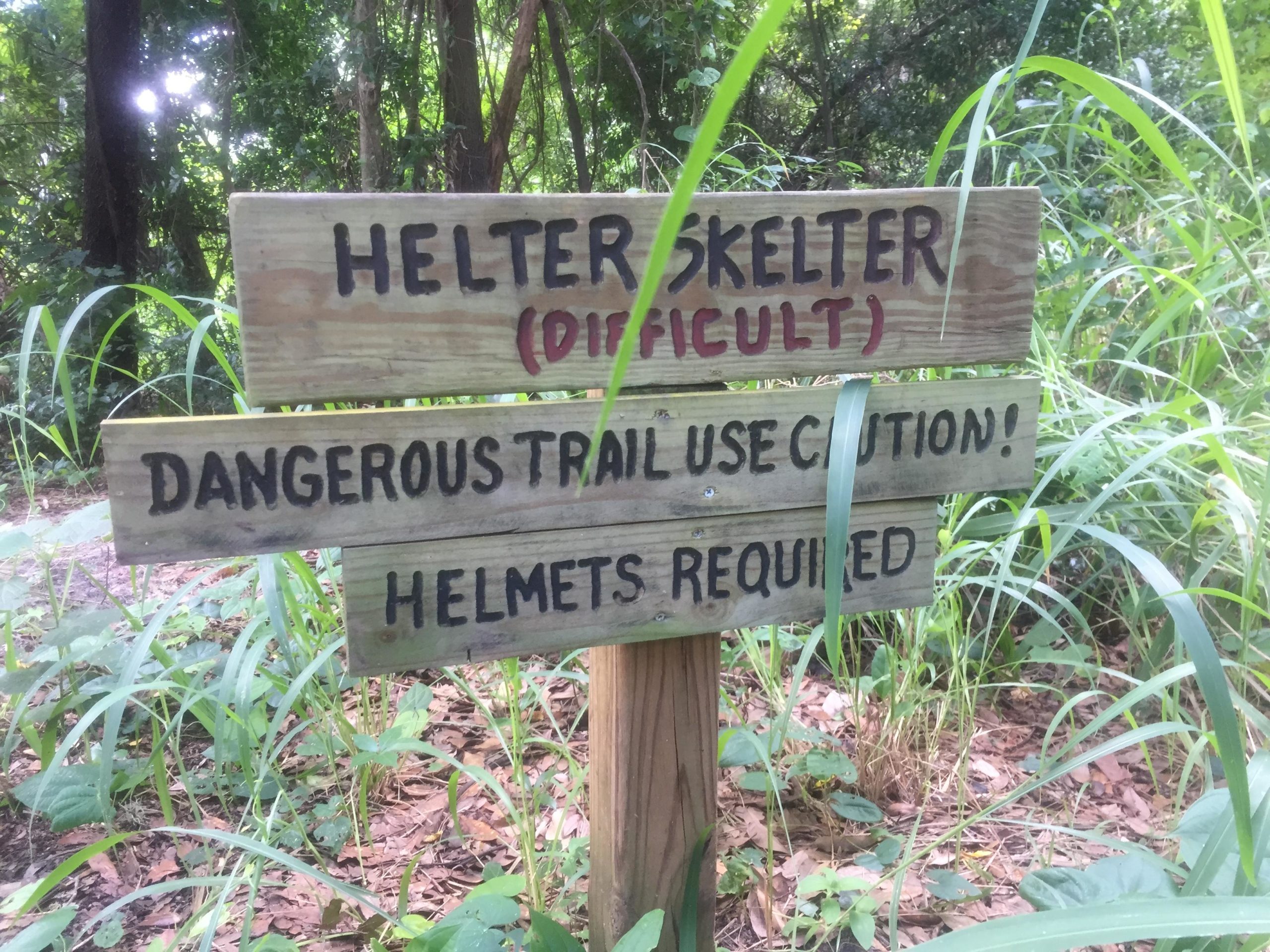 Wooden trail sign in a wooded area, reading "HELTER SKELTER (DIFFICULT)" at the top, followed by "DANGEROUS TRAIL USE CAUTION!" and "HELMETS REQUIRED," with tall grasses and foliage surrounding the sign. Helter Skelter mountain bike trail.