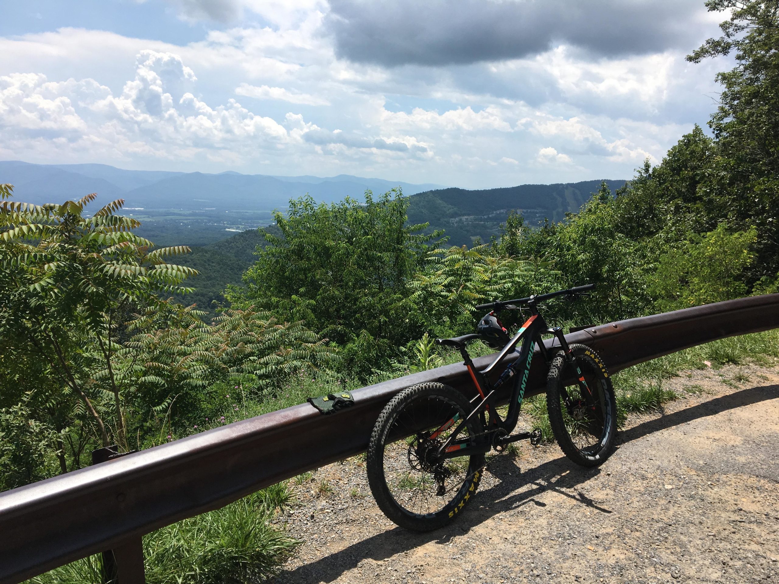A mountain bike resting against a guardrail, overlooking a scenic view of rolling hills and valleys under a partly cloudy sky. Lush greenery surrounds the area, adding to the natural landscape. Massanutten Western Slope mountain bike trail.