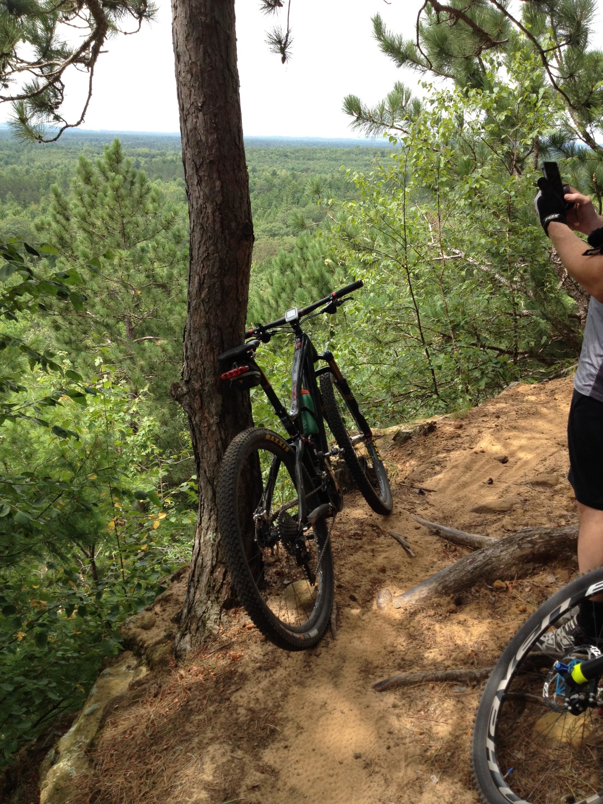 A mountain bike leaning against a tree on a sandy hillside, with a lush green landscape and distant trees in the background. A person is seen taking a photo nearby, wearing gloves and a short-sleeved shirt. The scene conveys an outdoor adventure atmosphere. Levis Mounds mountain bike trail.