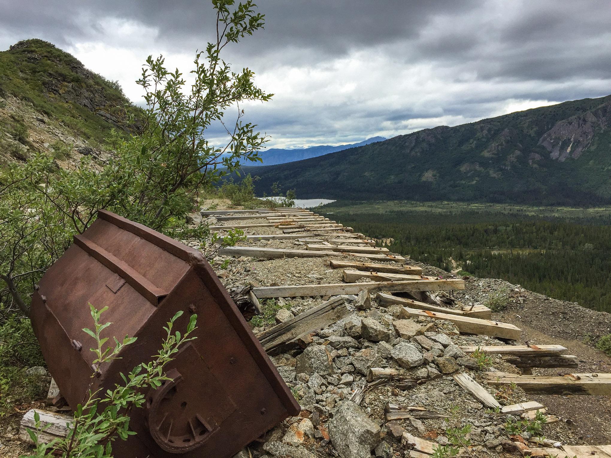 Landscape featuring a rusted metal object lying on its side, surrounded by greenery, with a view of a mountainous terrain and a river in the distance. The scene is under a cloudy sky, with wooden planks scattered along the ground. Atlin Mt SuperHero Trail mountain bike trail.