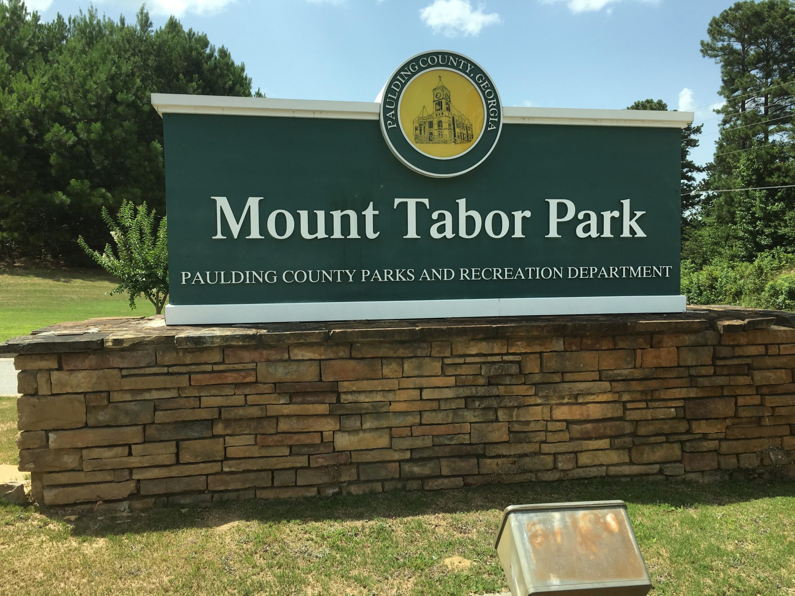 Sign for Mount Tabor Park, displayed on a stone base, with "Paulding County Parks and Recreation Department" written below. The background features greenery and clear blue skies. Mt Tabor Park mountain bike trail.