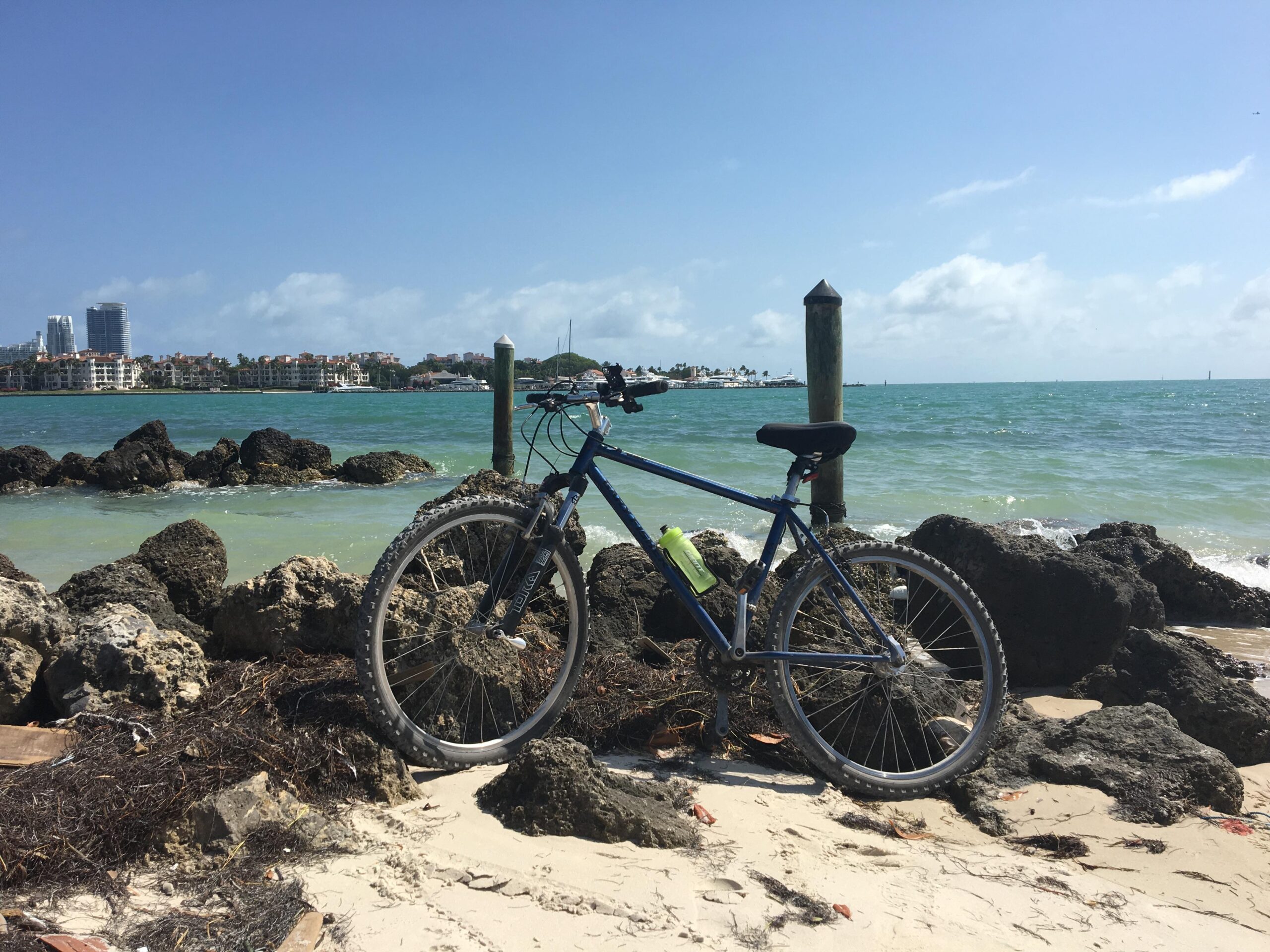 A blue mountain bike resting on rocks near a sandy beach, with a view of the turquoise sea and buildings in the background under a clear blue sky. Virginia Key North Point mountain bike trail.