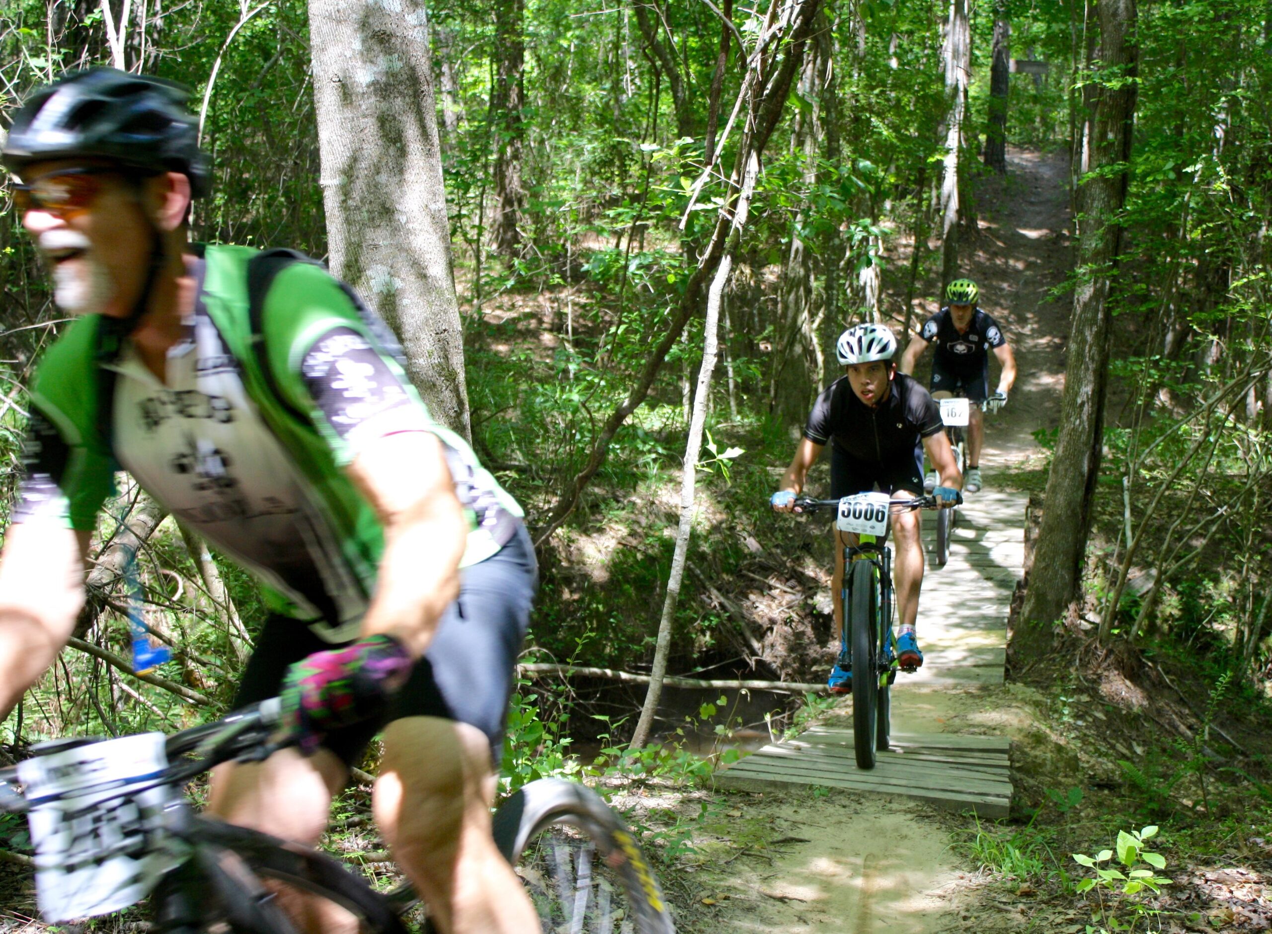 Three mountain bikers navigate a narrow trail through a lush, green forest. One cyclist in the foreground is blurred due to movement, while the others are on a wooden bridge over a small creek in the background. Sunlight filters through the trees, highlighting the vibrant foliage surrounding the path. Mt. Zion Bike Trails mountain bike trail.