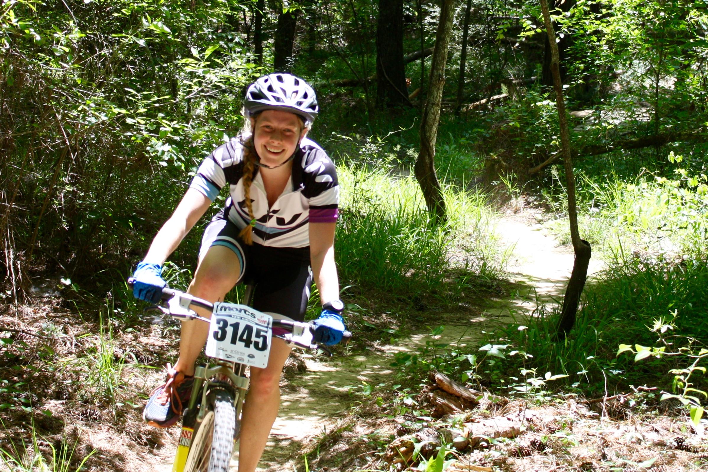 A young female mountain biker rides along a dirt path in a wooded area, wearing a helmet and a cycling jersey. She smiles as she navigates the trail, surrounded by greenery and trees. Mt. Zion Bike Trails mountain bike trail.