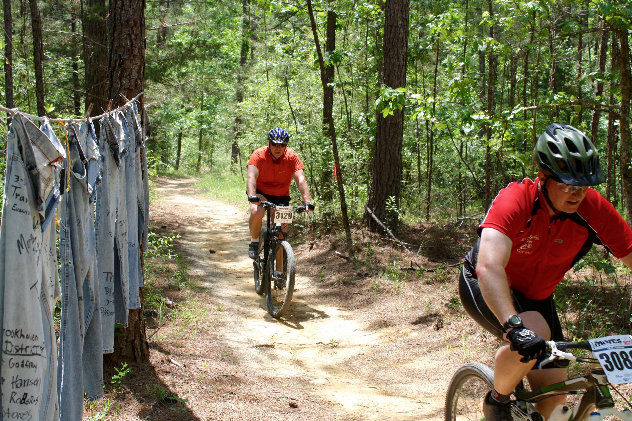 Two mountain bikers navigate a dirt trail surrounded by trees. To the left, a row of denim jeans is hung on a line, each marked with messages. The cyclists are wearing helmets and numbered race bibs, with one in an orange shirt and the other in a red shirt. The sun shines through the foliage, creating a vibrant outdoor scene. Mt. Zion Bike Trails mountain bike trail.