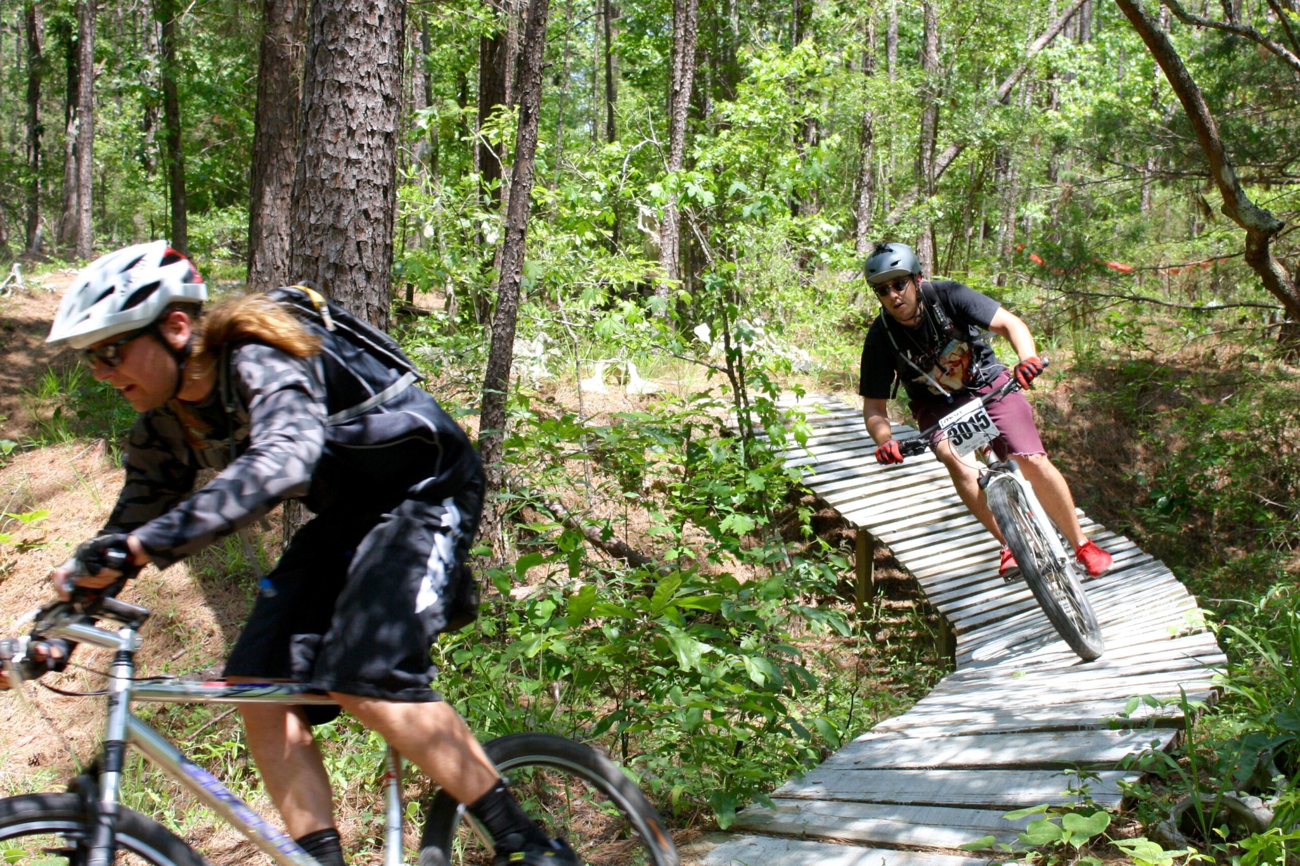Two mountain bikers navigating a wooden bridge on a forest trail. One rider, wearing a helmet and sunglasses, is leaning forward on their bike, while the other rider is approaching the bridge, wearing a black shirt and red shorts. The scene is surrounded by lush greenery and tall trees, highlighting an outdoor activity in a natural setting. Mt. Zion Bike Trails mountain bike trail.
