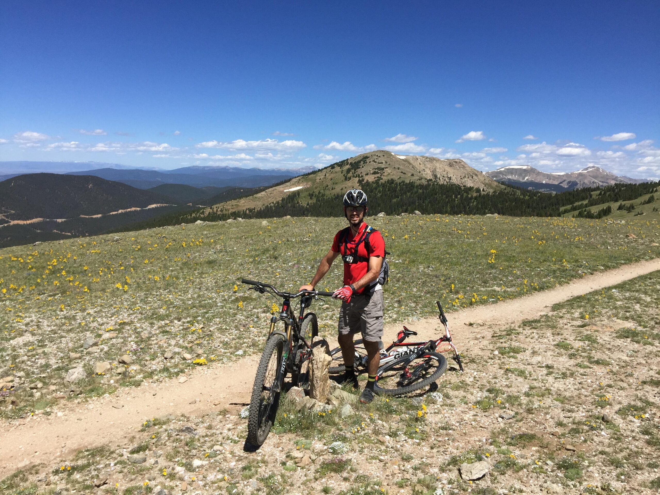 A mountain biker stands next to his bike on a dirt trail in a grassy, flower-covered landscape. The background features rolling hills and distant mountains under a clear blue sky. Monarch Crest Trail mountain bike trail.