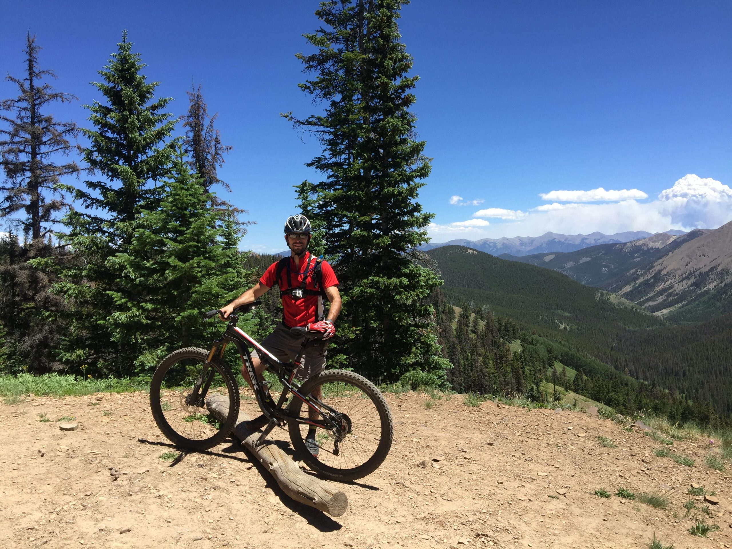 A mountain biker in a red shirt and helmet stands next to his mountain bike on a dirt trail surrounded by green pine trees. In the background, there are rolling hills and mountains under a clear blue sky with a few clouds. Monarch Crest Trail mountain bike trail.