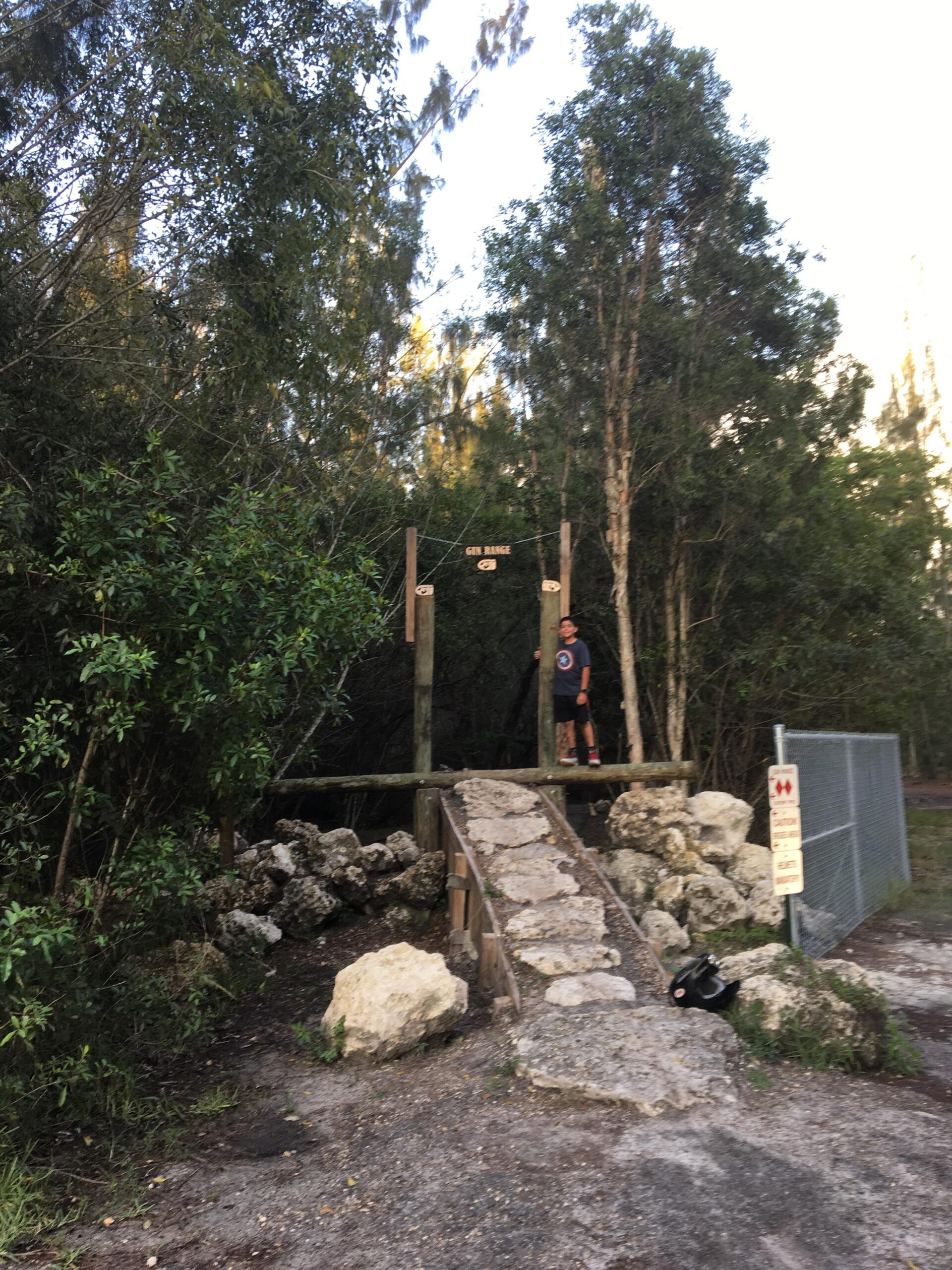A person standing on a wooden platform at the entrance of a gun range, surrounded by trees and greenery. A rocky pathway leads to the platform, with a fence and a caution sign visible nearby. Markham Park mountain bike trail.