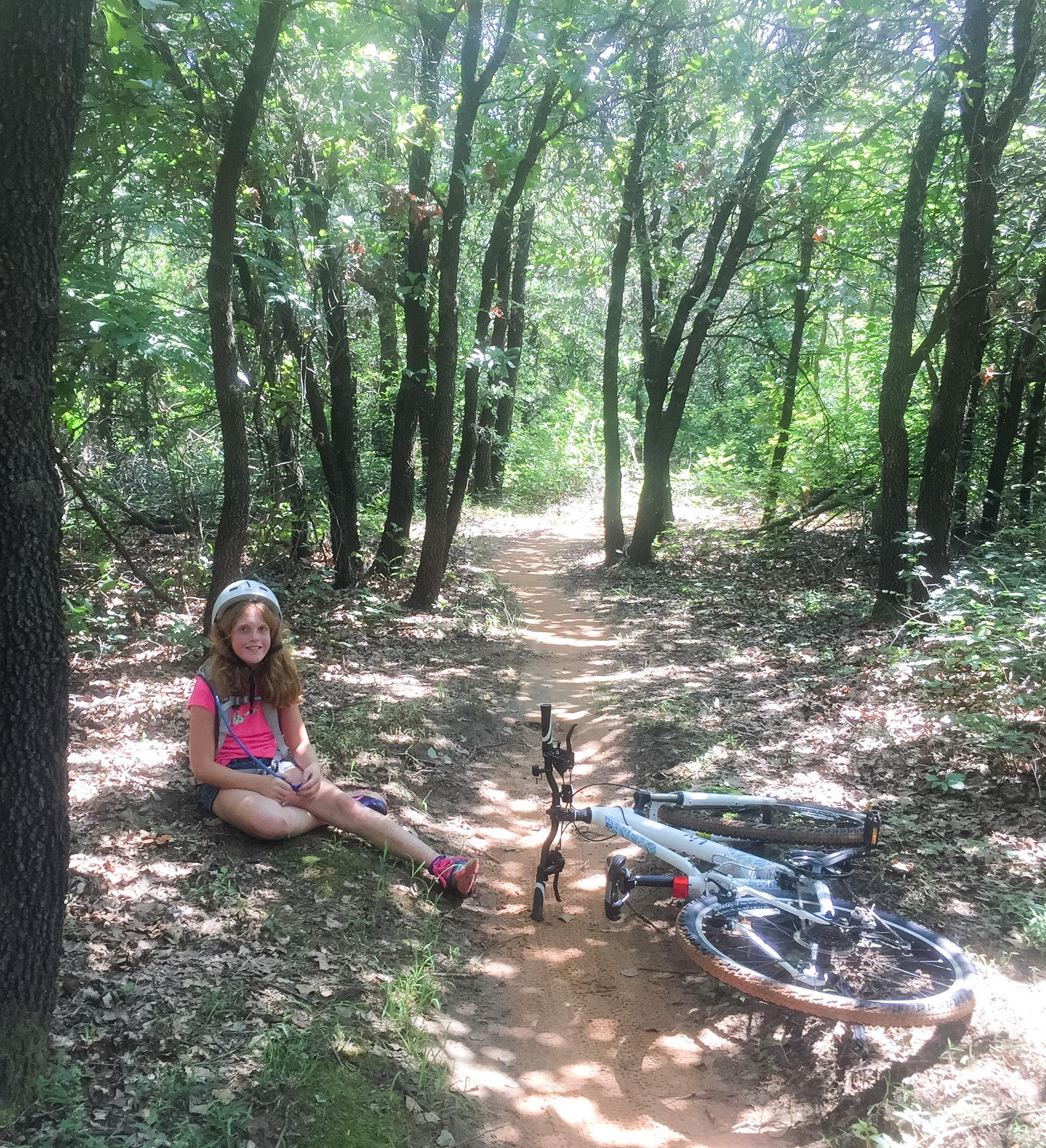 A young girl wearing a helmet sits on the ground beside a mountain bike, surrounded by a lush forest with sunlight filtering through the trees. A dirt trail winds through the background, and fallen leaves cover the ground. The scene conveys a sense of adventure and outdoor enjoyment. Lake Stanley Draper mountain bike trail.