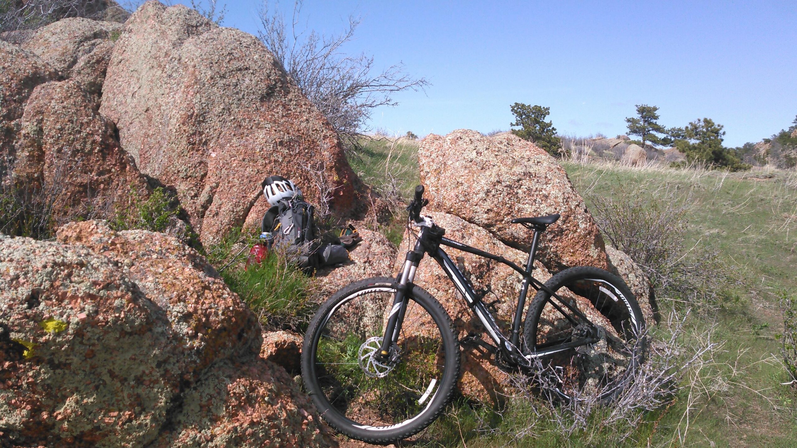 Specialized Rockhopper Comp 29: A mountain bike leaning against a large rock formation in a grassy landscape, with a helmet and backpack resting nearby under a clear blue sky.