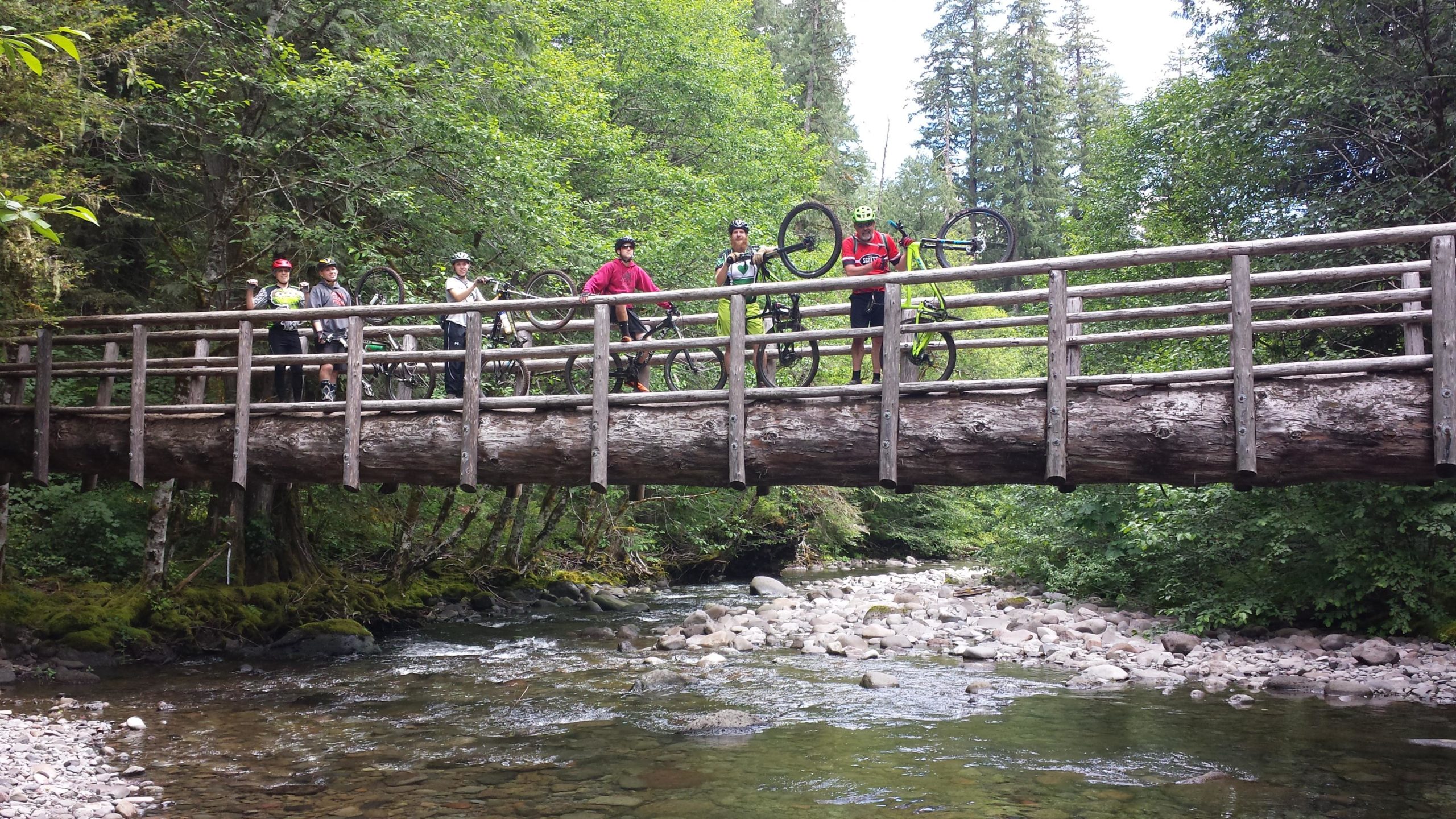 A group of six mountain bikers stands on a wooden bridge over a clear, rocky river. They are surrounded by lush green trees and foliage. Each biker is holding a mountain bike, with varying colors of helmets and clothing. The atmosphere is lively and adventurous, capturing a moment of outdoor fun. Mckenzie River Trail mountain bike trail.