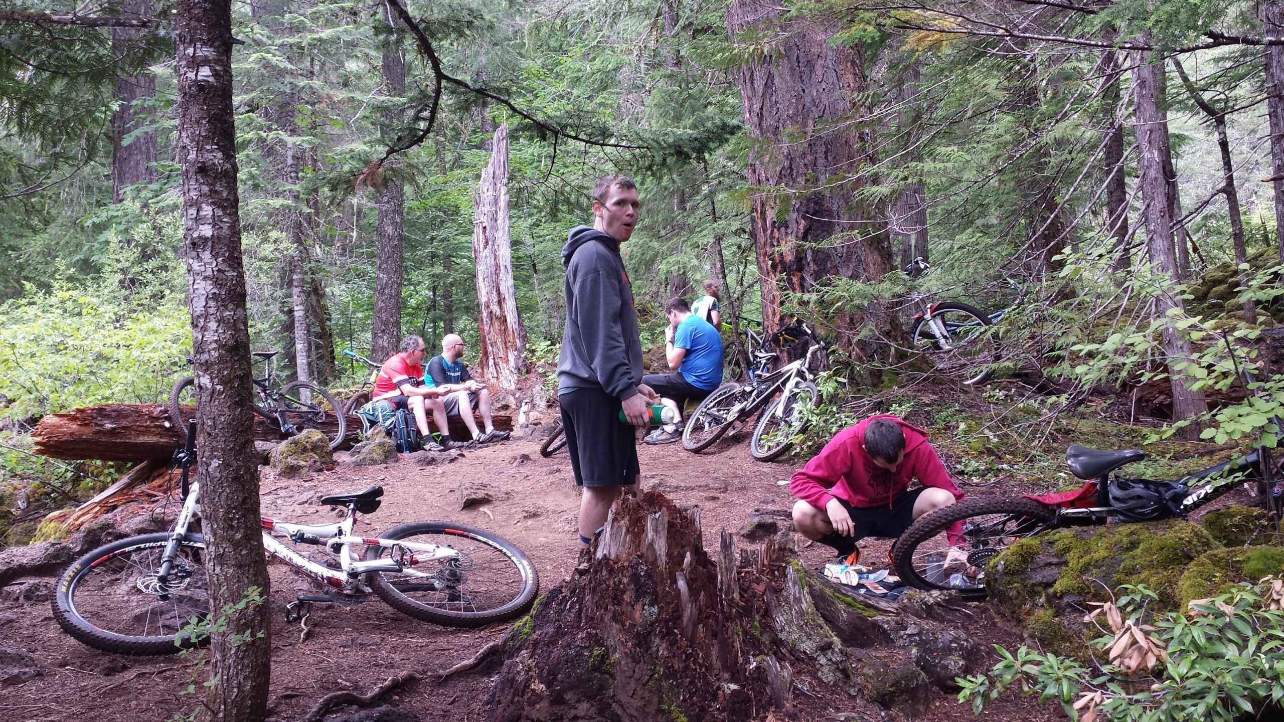 A group of mountain bikers resting in a forested area, surrounded by trees and rocky terrain. Some individuals are seated on logs, while others are tending to their bikes on the ground. Bicycles are scattered around, and the atmosphere is casual and relaxed. Mckenzie River Trail mountain bike trail.