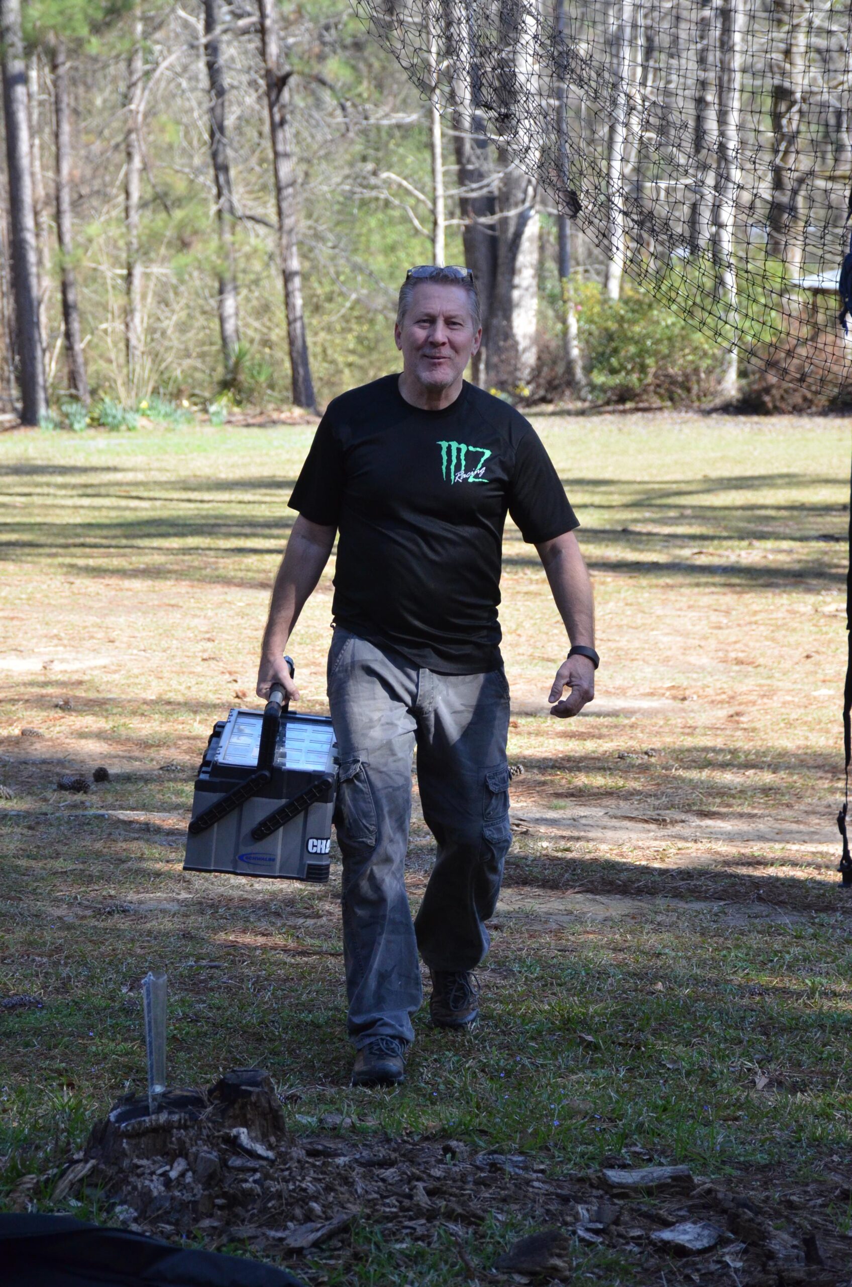 A man in a black t-shirt and grey cargo pants carrying a toolbox walks across a grassy area surrounded by trees. Sunlight filters through the foliage, creating dappled shadows on the ground. Mt. Zion Bike Trails mountain bike trail.