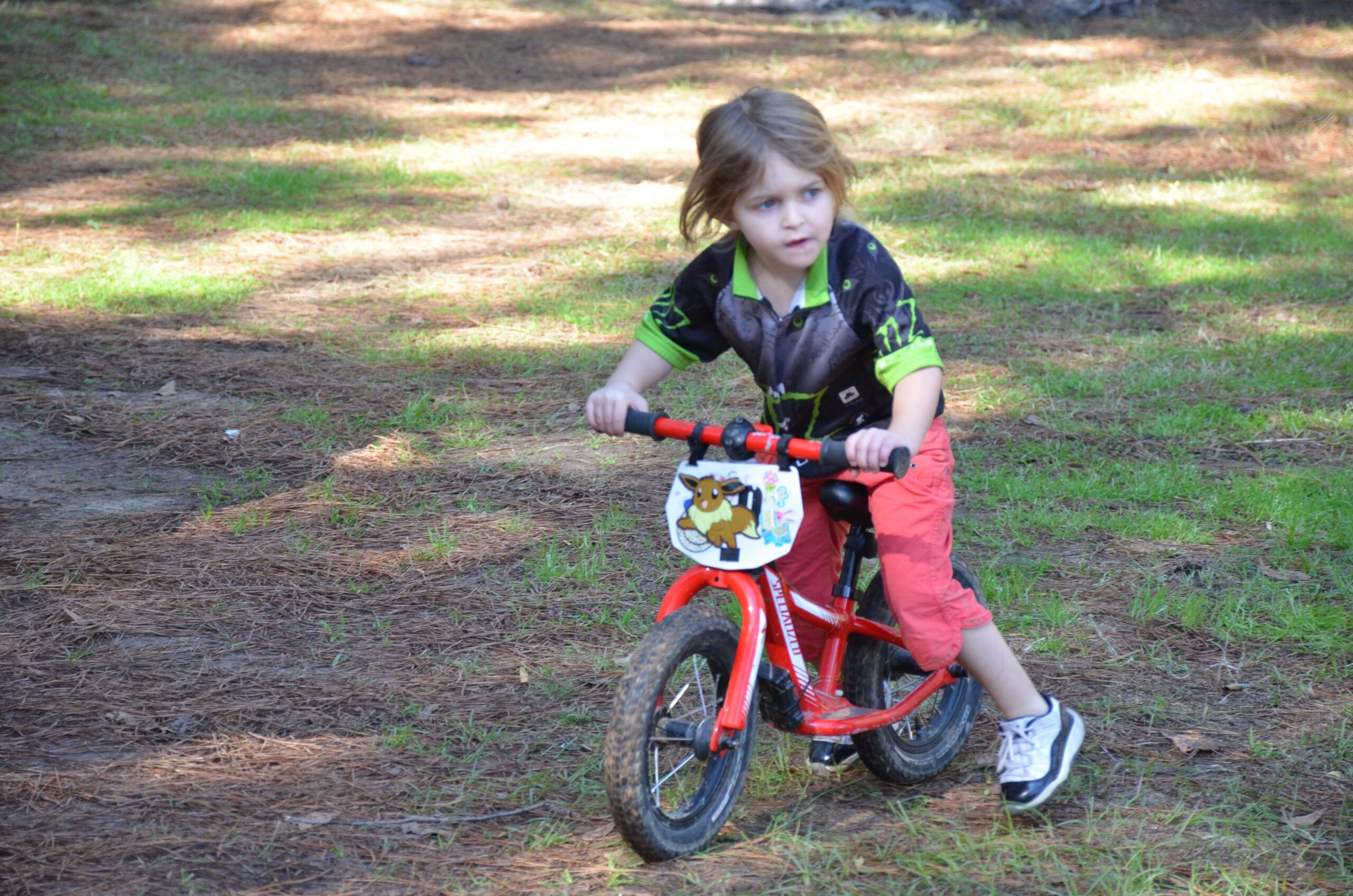 A young child riding a small red bike on a trail covered with pine needles and green grass. The child is wearing a black and green shirt and red shorts, and appears focused while biking. Mt. Zion Bike Trails mountain bike trail.