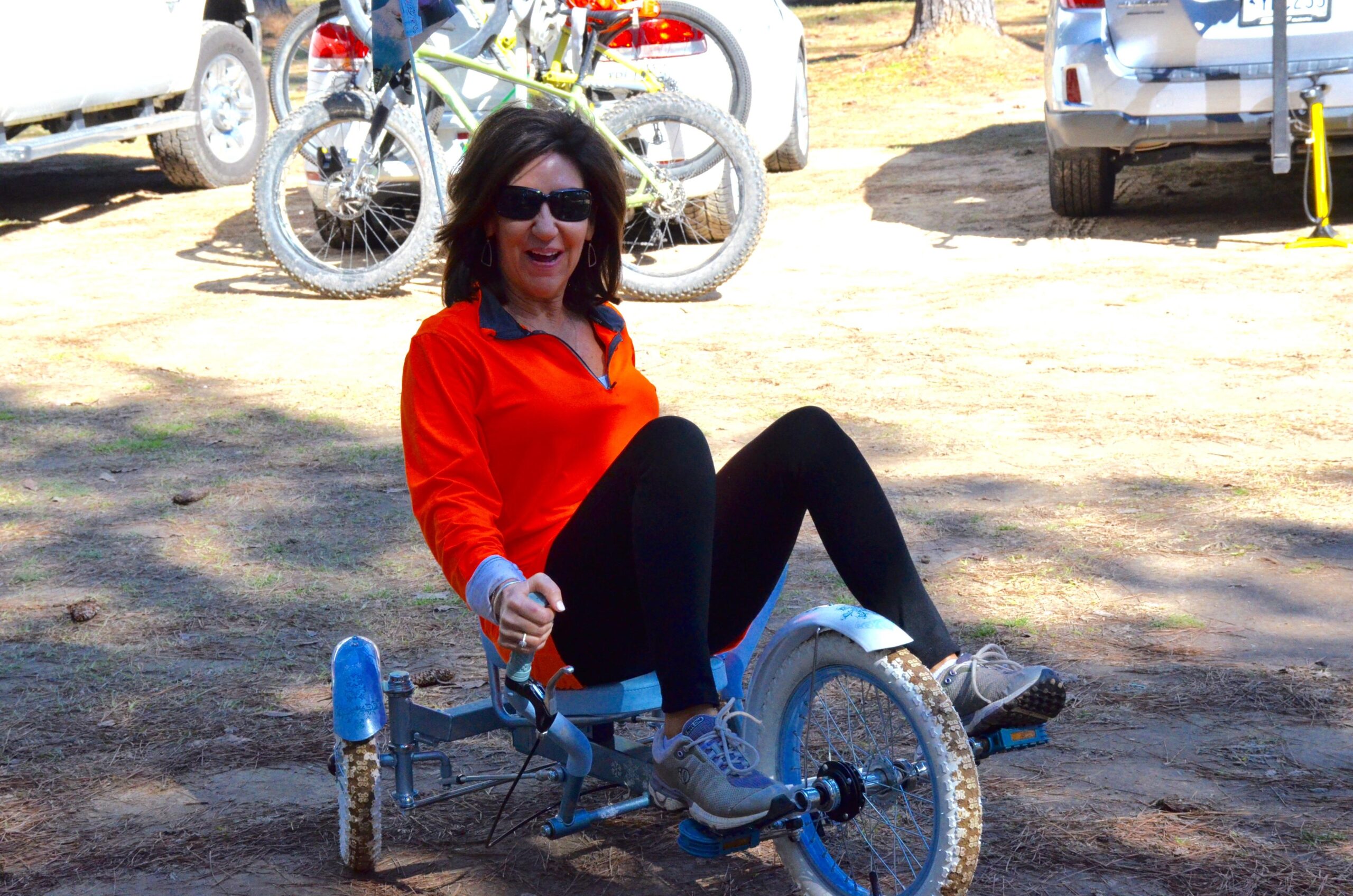 A woman wearing an orange long-sleeve shirt and black leggings is sitting on a three-wheeled pedal cart in a natural outdoor setting. She is smiling and appears to be enjoying herself, with her legs bent and feet positioned on the pedals. In the background, there are bicycles and parked vehicles, indicating an active outdoor environment. Mt. Zion Bike Trails mountain bike trail.