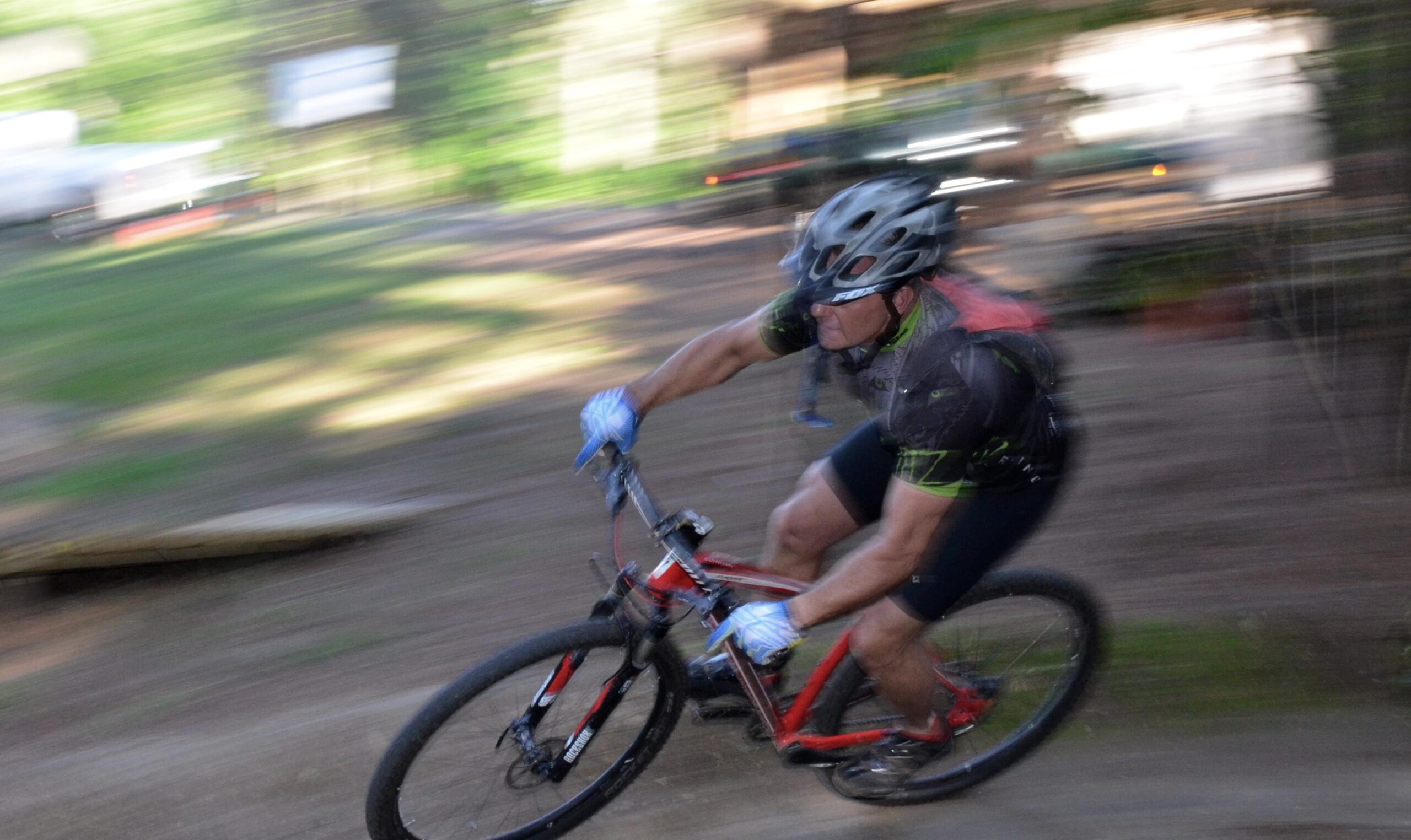 A mountain biker in motion, leaning into a turn on a dirt trail, surrounded by greenery. The bike is red and the rider is wearing a helmet and blue gloves, indicating a sporty and adventurous atmosphere. The image captures a sense of speed and movement with a blurred background. Mt. Zion Bike Trails mountain bike trail.