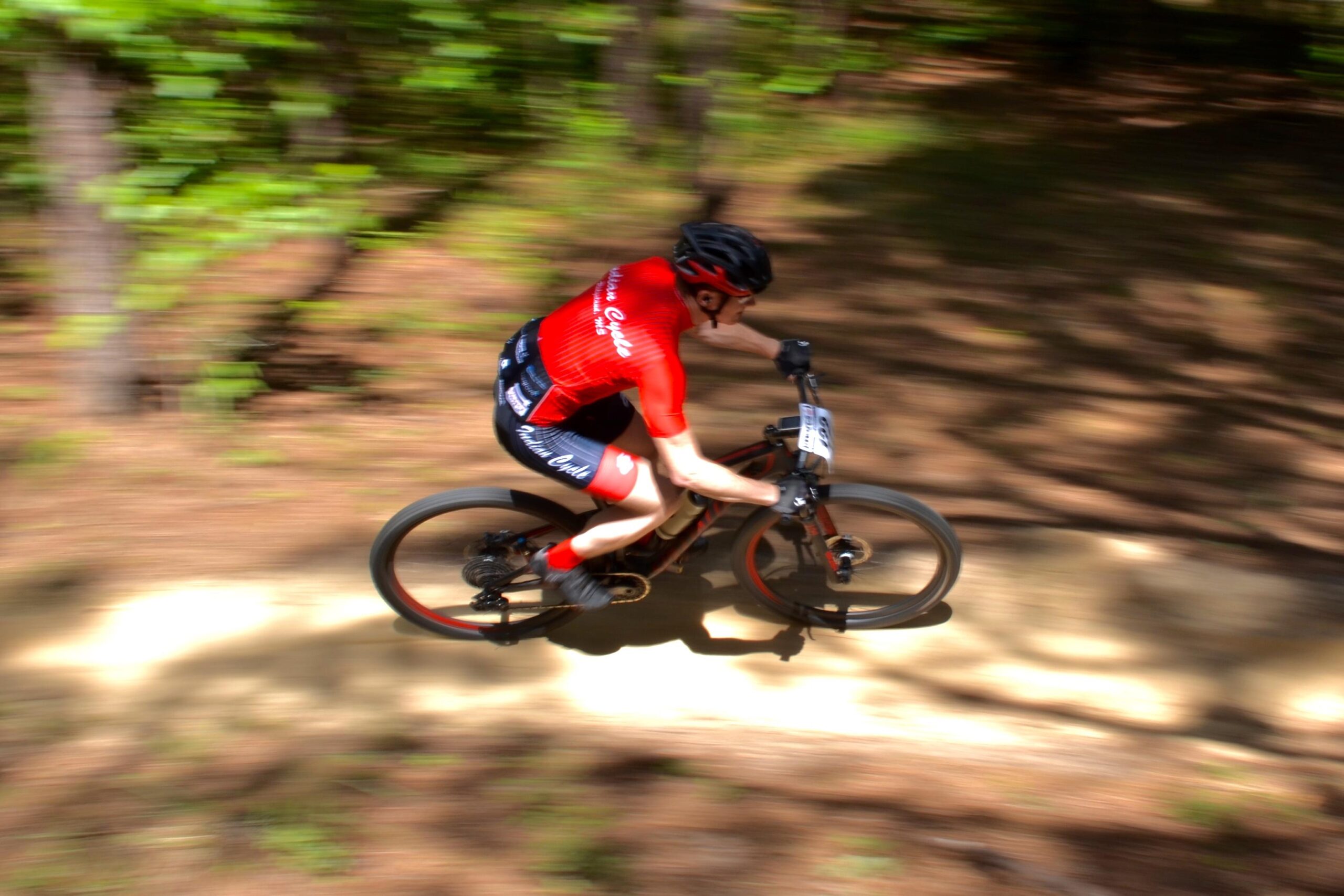 A cyclist wearing a red jersey and helmet speeds through a wooded trail, capturing a sense of motion with a blurred background. The cyclist is focused on the path ahead, with trees and greenery surrounding the trail. Mt. Zion Bike Trails mountain bike trail.