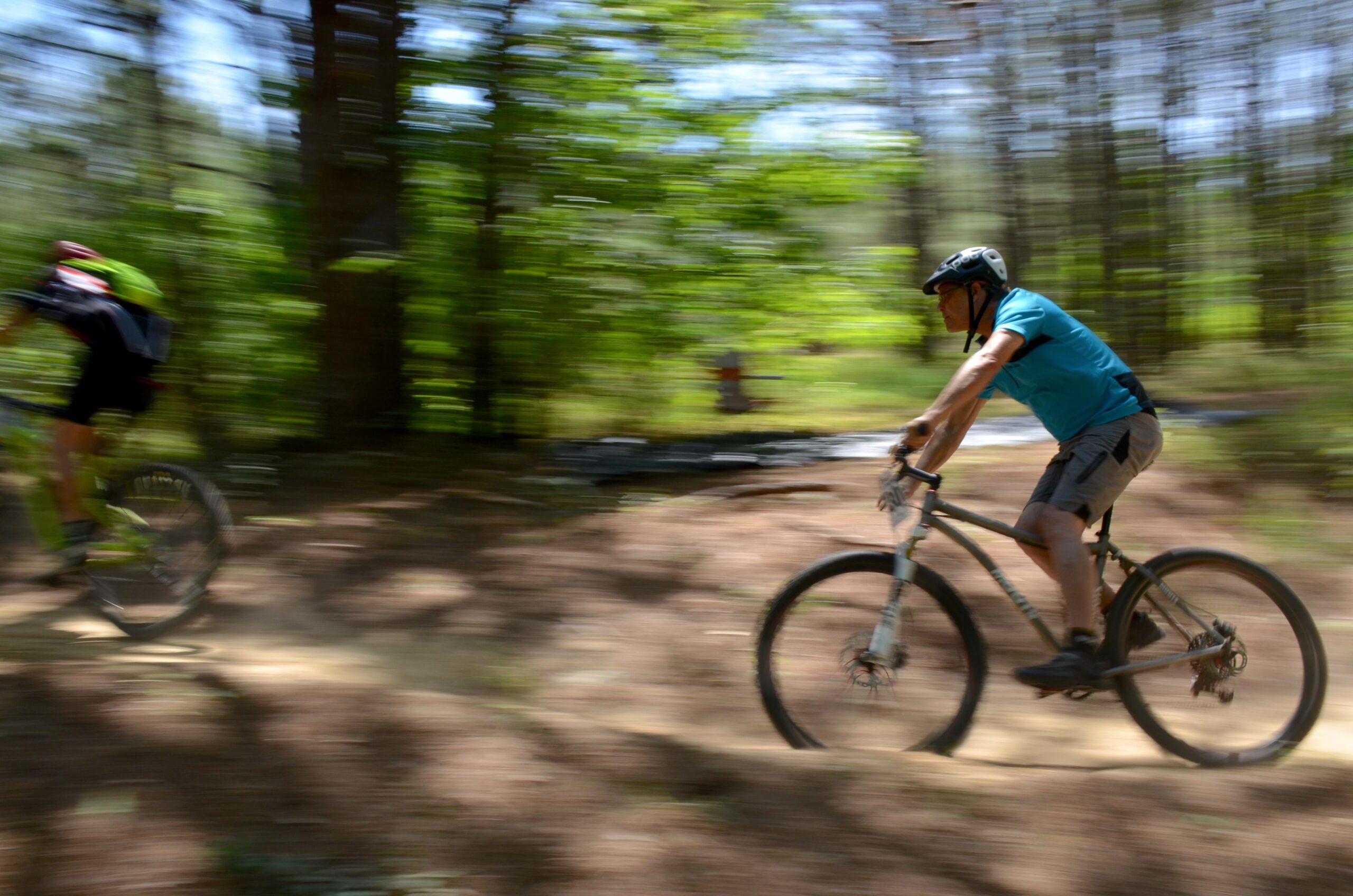 Two mountain bikers riding along a wooded trail, with one rider in focus and the other slightly blurred, suggesting motion. The scene is filled with green foliage and sunlight filtering through the trees, enhancing the sense of speed and action. Mt. Zion Bike Trails mountain bike trail.