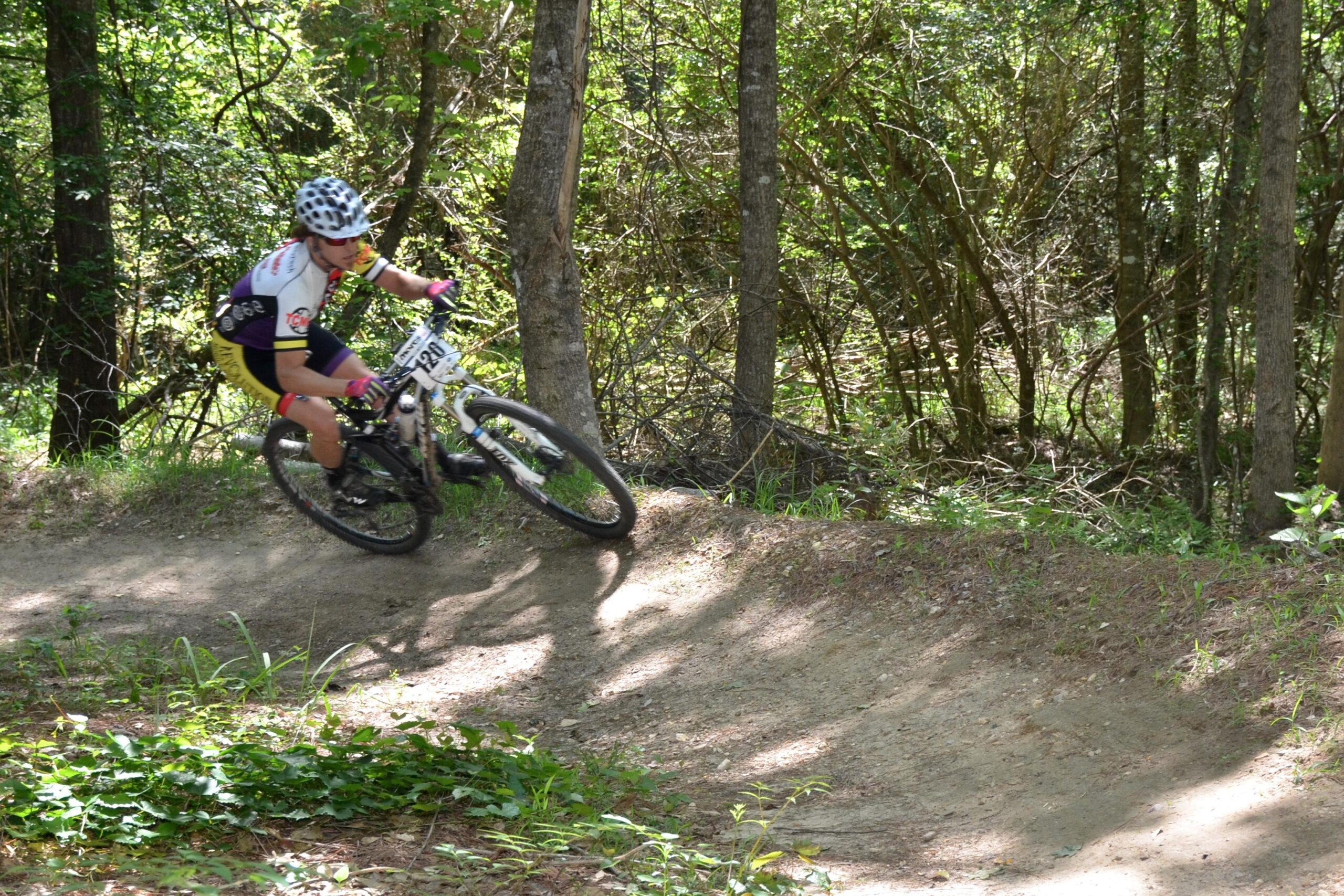 A cyclist in a helmet and colorful biking gear leans into a turn on a dirt trail surrounded by trees. Sunlight filters through the foliage, casting dappled shadows on the ground as the rider maneuvers expertly around the curve. Mt. Zion Bike Trails mountain bike trail.