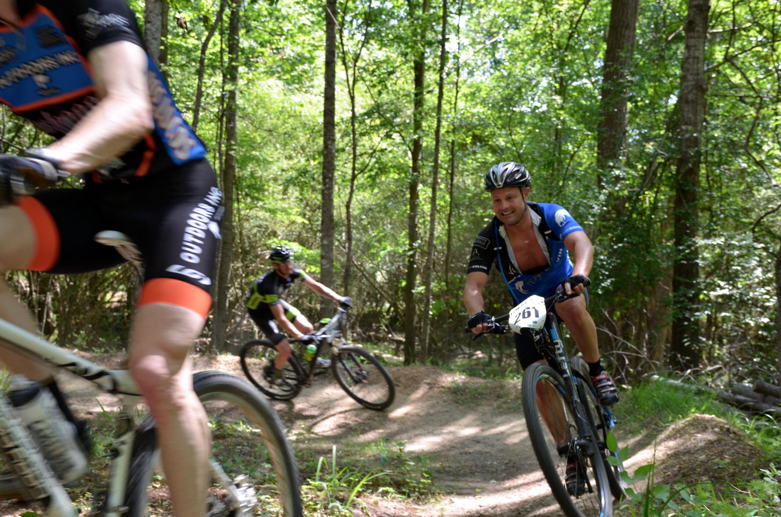 A group of three mountain bikers navigating a forested trail on a sunny day. The foreground features a rider in an orange and black uniform, while another bike can be seen slightly blurred in the background. The third cyclist, wearing a blue jersey with the number 261, is smiling as he rides along the curved path surrounded by lush greenery. Mt. Zion Bike Trails mountain bike trail.