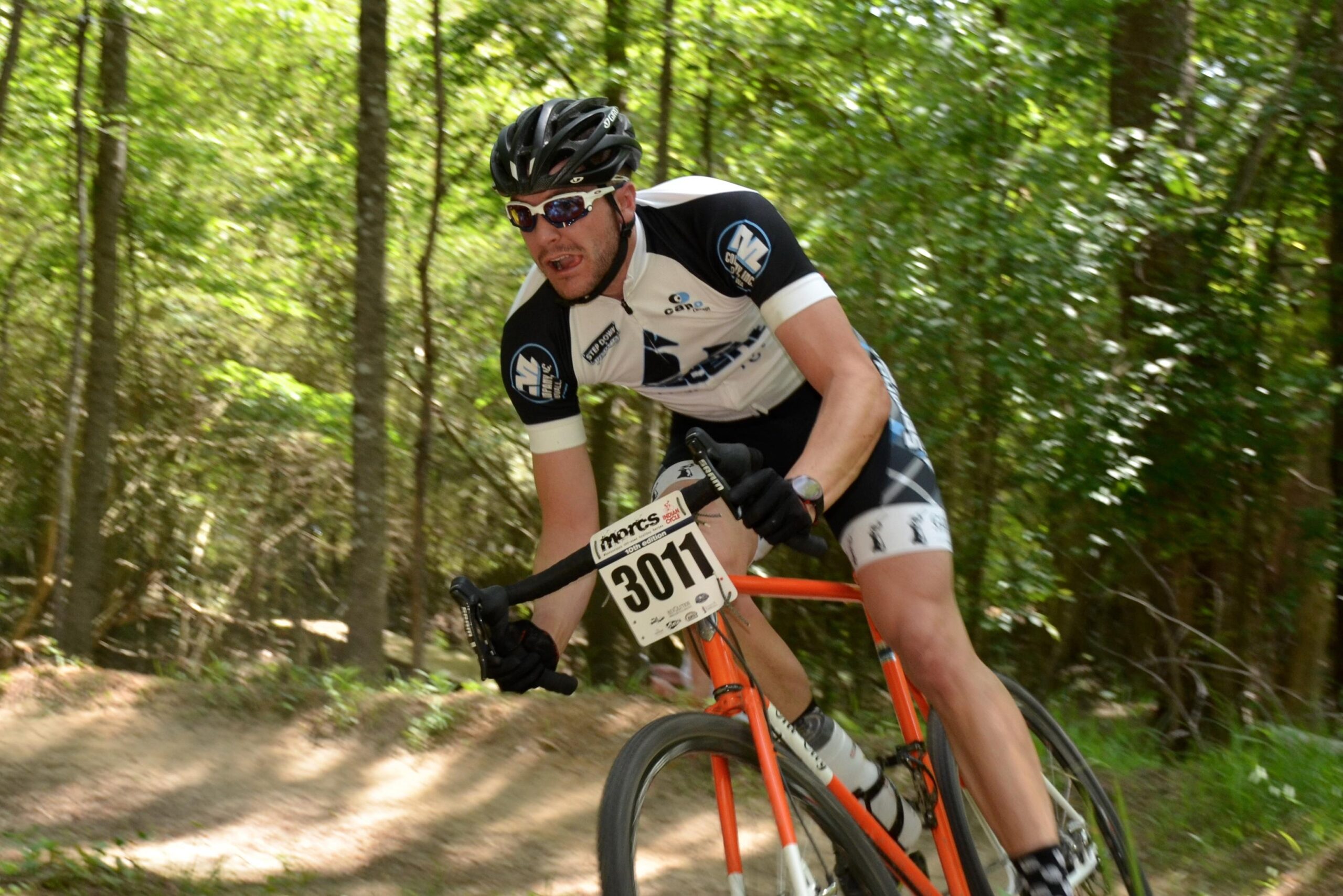 A mountain biker in a black and white cycling jersey and helmet rides swiftly on a forested trail. The bike is orange, and a race number (3011) is visible on the front. The surroundings are lush with green trees, creating a vibrant outdoor setting. Mt. Zion Bike Trails mountain bike trail.