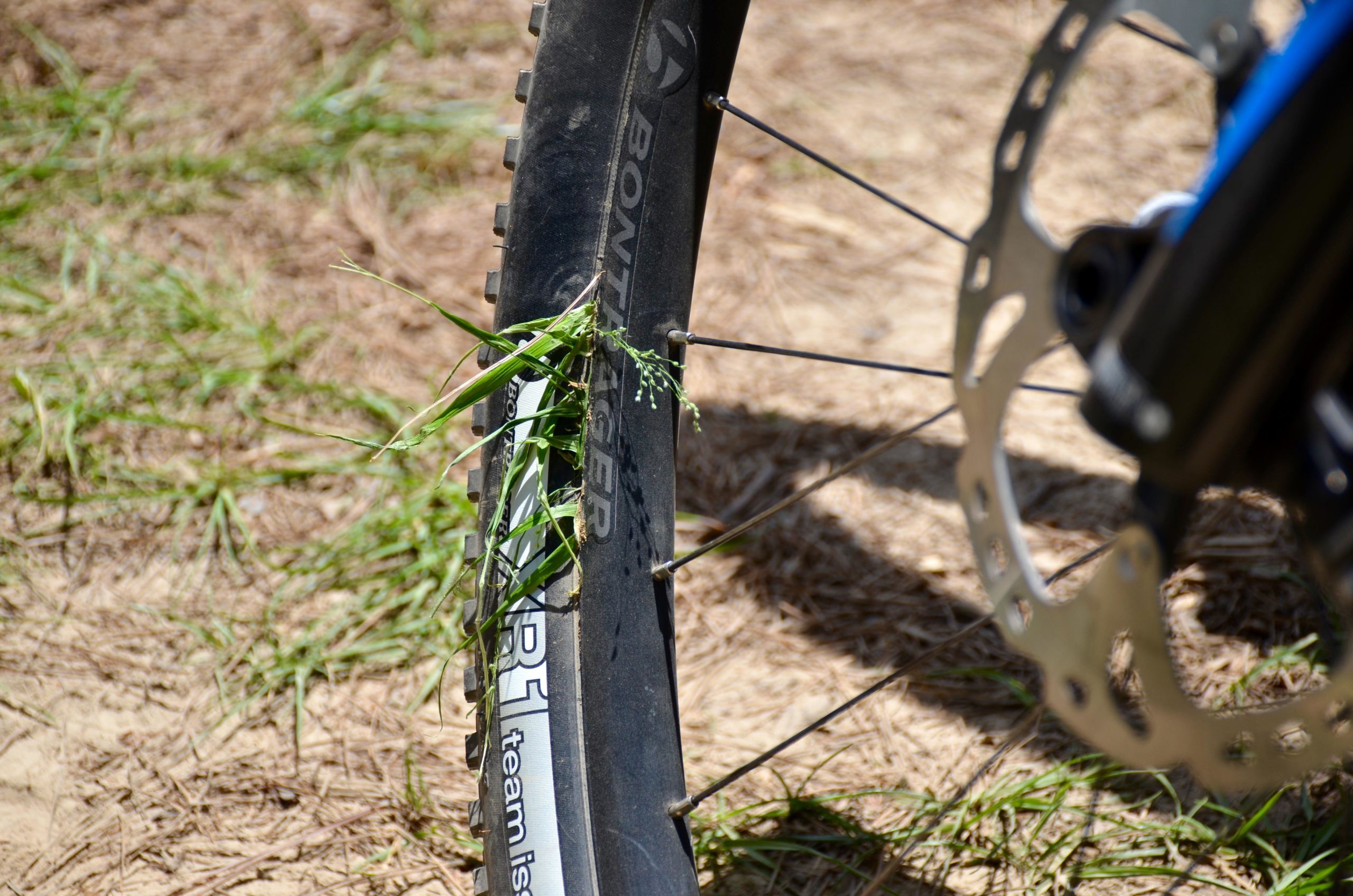 A close-up view of a bicycle tire with grass and weeds stuck in it, set against a background of dirt and grass. The tire is labeled with the brand name "Bontrager" and shows signs of outdoor use. Mt. Zion Bike Trails mountain bike trail.