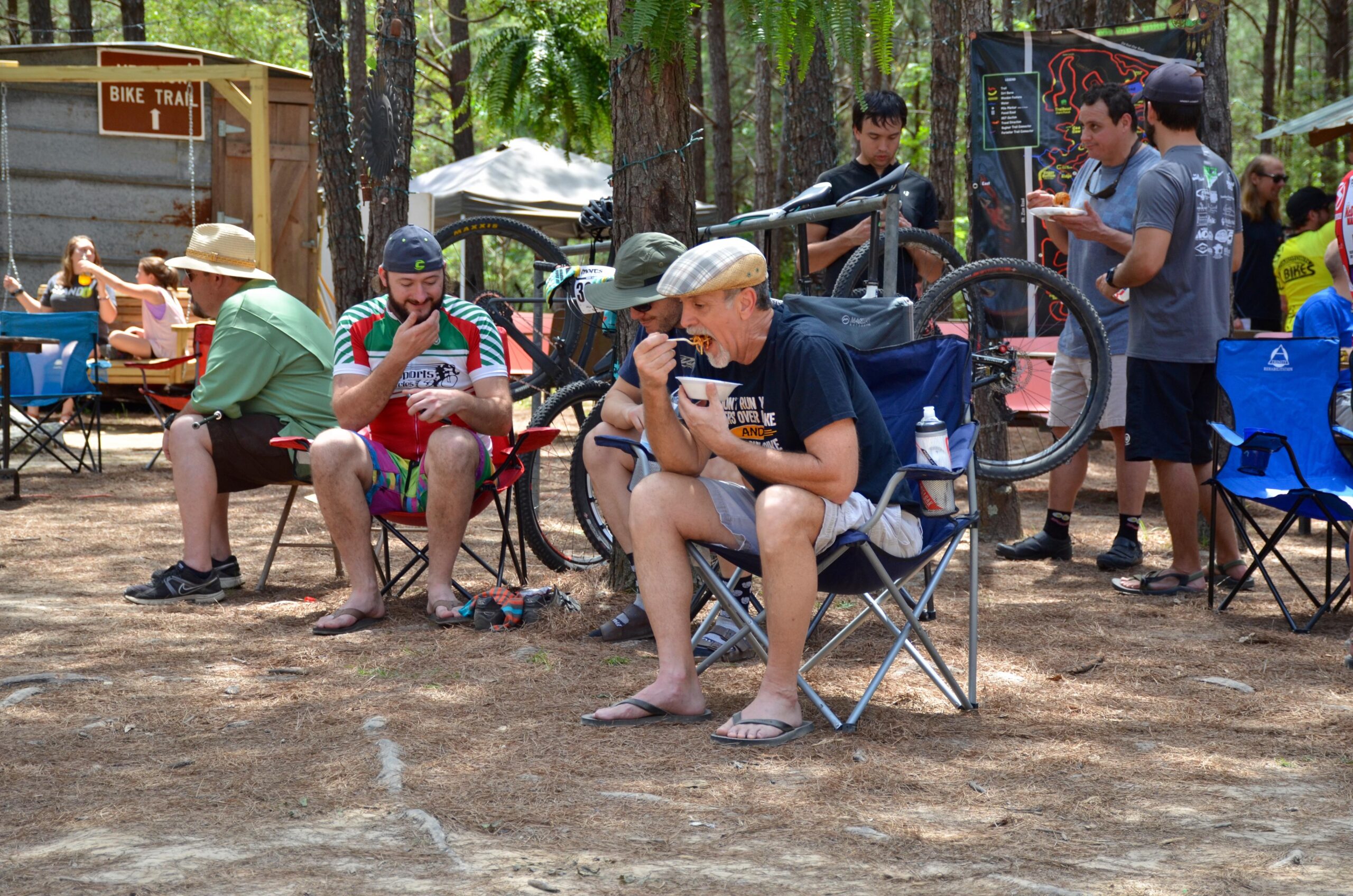 A group of people relaxing outdoors in a wooded area, some sitting on camping chairs. A man wearing a red and green cycling jersey eats from a bowl, while another man in a hat enjoys food from his hand. In the background, others are gathered, with bicycles visible and a sign pointing towards a bike trail. The scene conveys a casual, outdoor gathering atmosphere. Mt. Zion Bike Trails mountain bike trail.