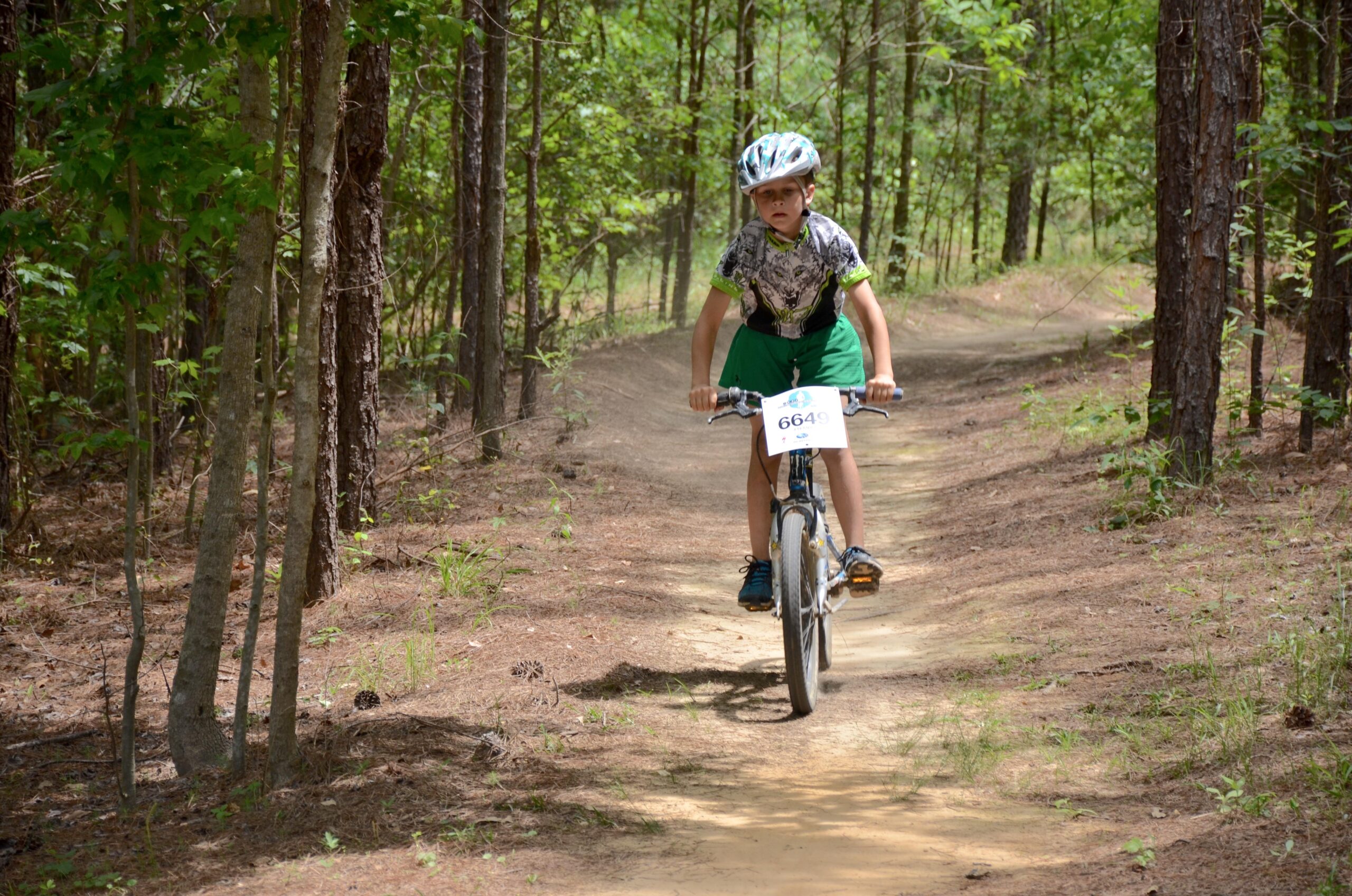 A young boy riding a bicycle on a dirt trail in a wooded area. He is wearing a helmet and a colorful shirt, with a race number attached to his bike. The background features trees and greenery, indicating a natural outdoor setting. Mt. Zion Bike Trails mountain bike trail.