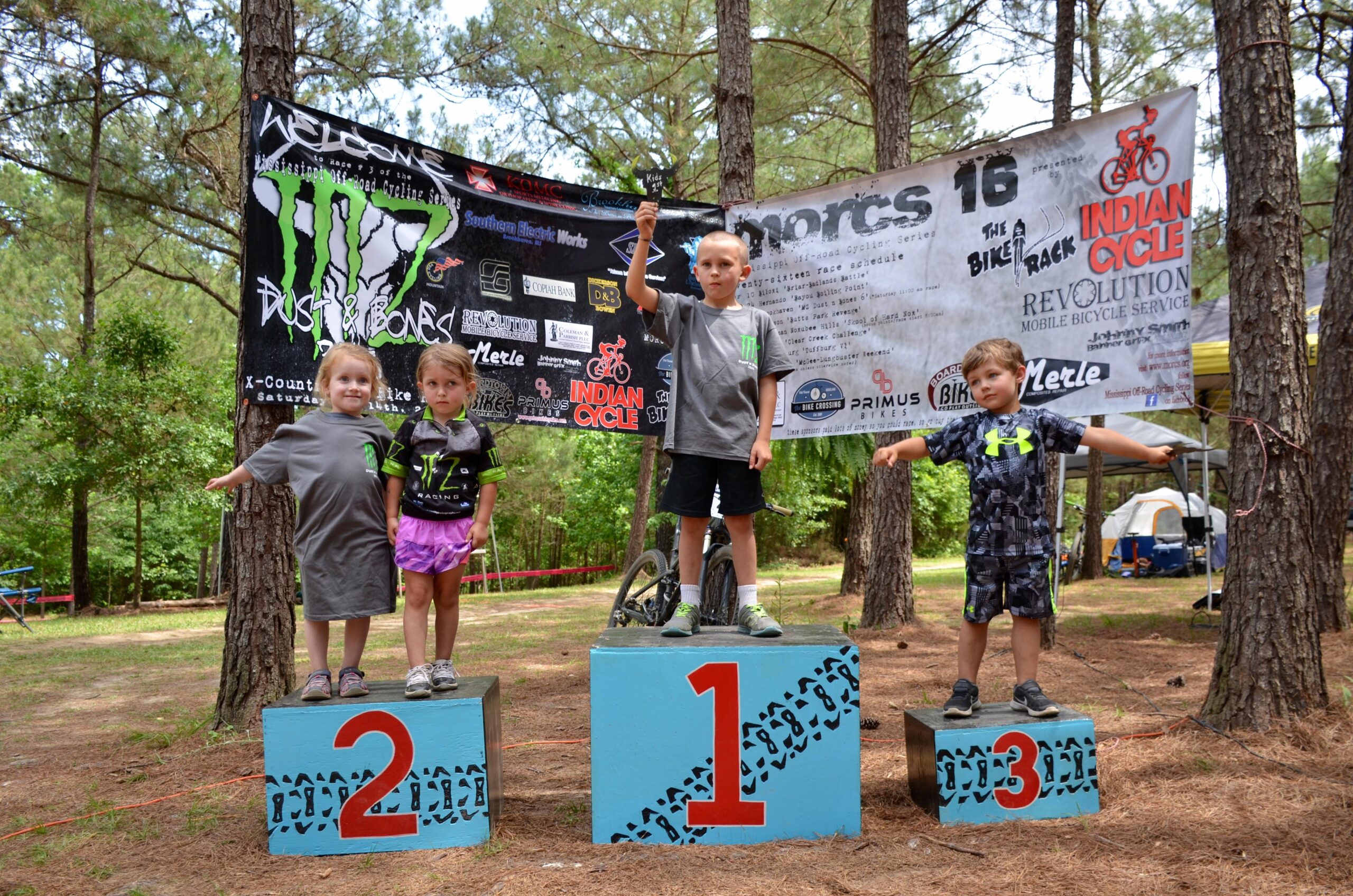 Four children stand on podiums at an outdoor event celebrating their achievements. The child on the first podium, wearing a gray shirt and holding a trophy, looks proudly at the camera. The child on the second podium is dressed in a Monster Energy shirt, while another child in a pink outfit stands next to her. The child on the third podium, wearing a black and green outfit, poses with arms outstretched. In the background, a banner with event details and trees can be seen, creating a lively and festive atmosphere. Mt. Zion Bike Trails mountain bike trail.