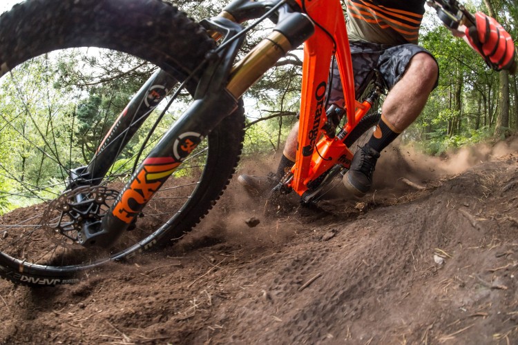 A close-up view of an orange mountain bike navigating a dirt trail, with dust being kicked up from the rear wheel. The rider, wearing dark shorts and black shoes, leans into a turn, showcasing the bike's front suspension and tire treads. The surrounding forest greenery is softly blurred, emphasizing the action and movement.