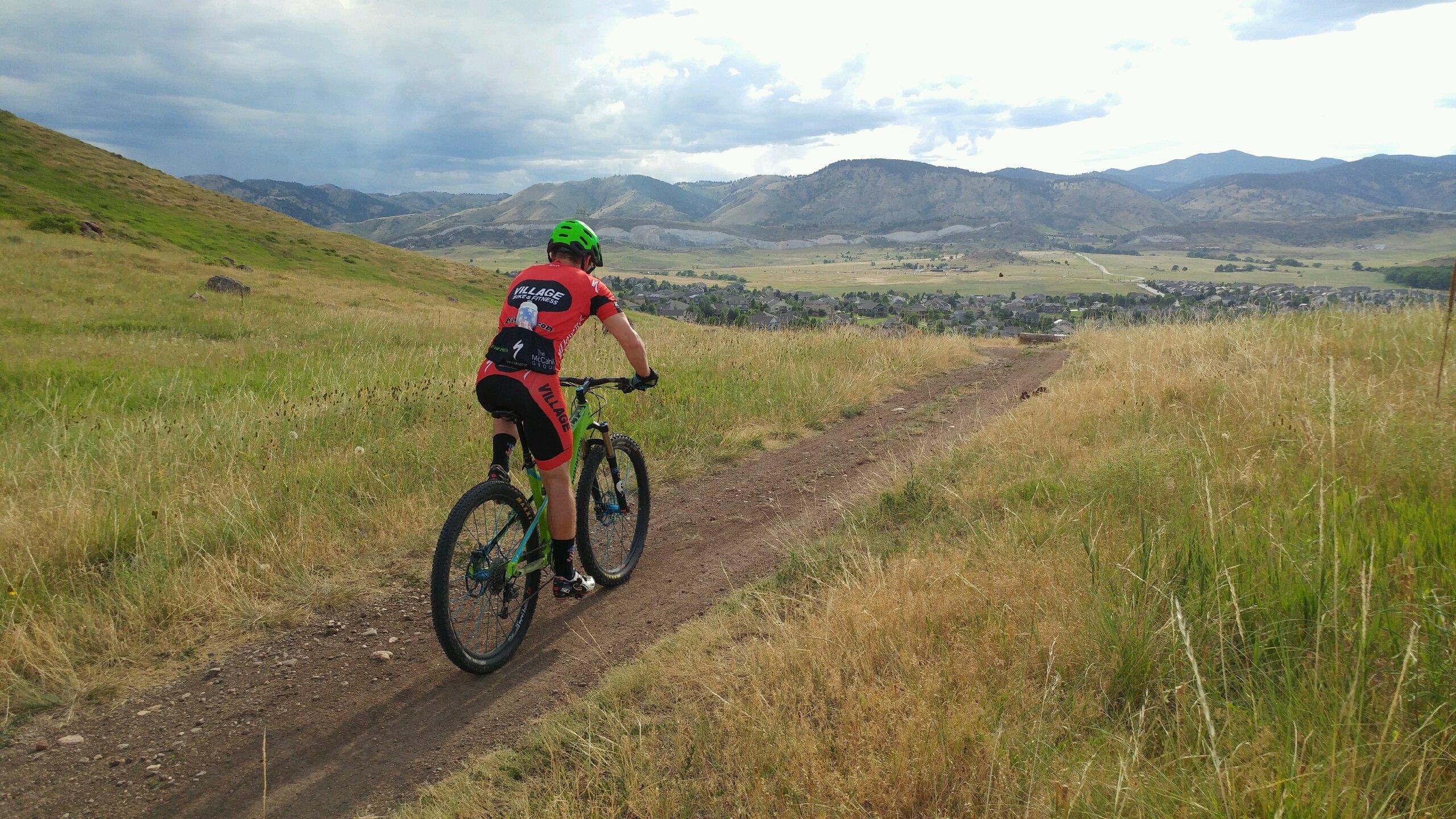 A cyclist wearing a red and black jersey rides a mountain bike along a dirt trail surrounded by tall grass, with rolling hills and mountains in the background under a cloudy sky. North Table Mountain mountain bike trail.