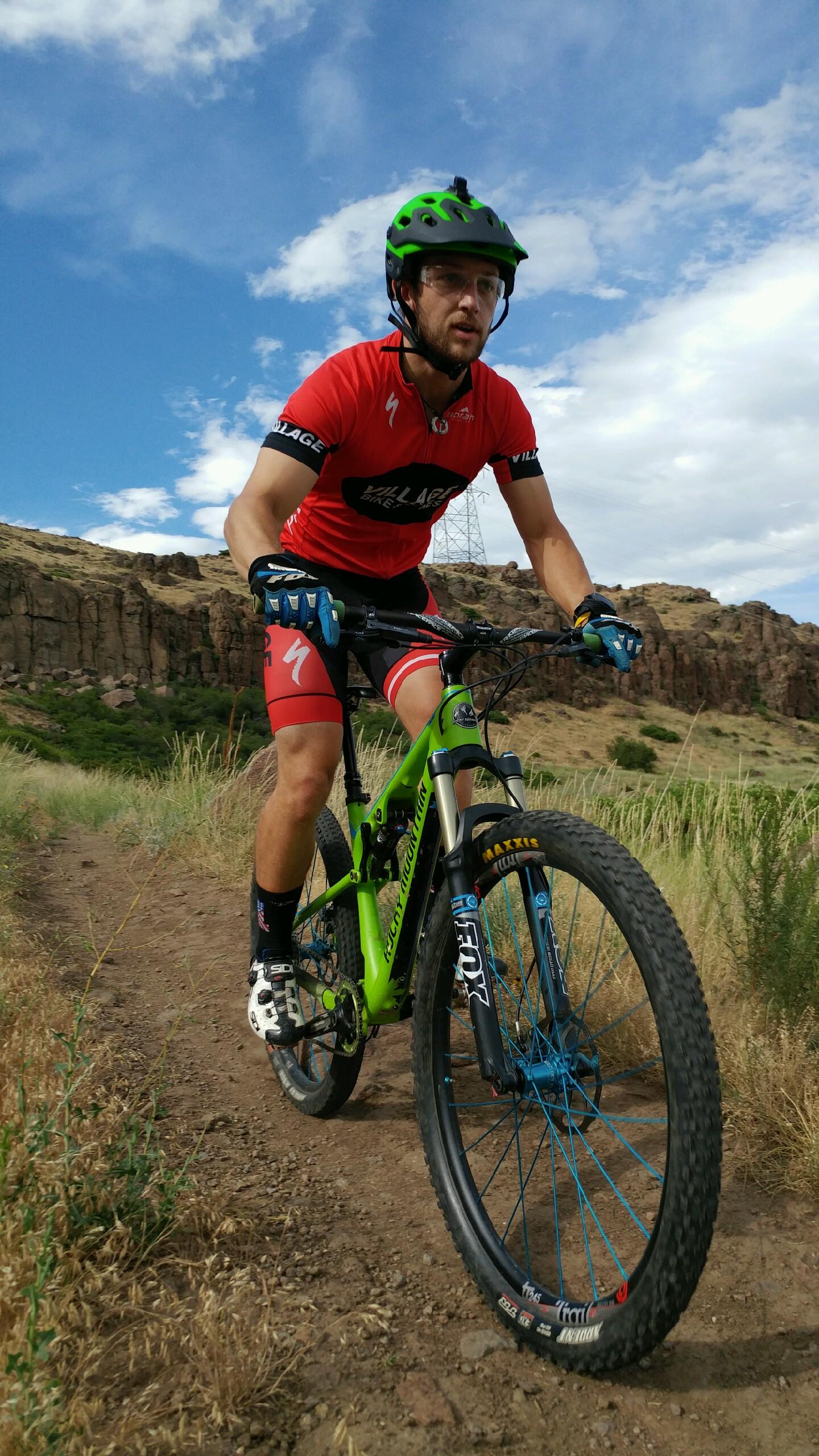 A mountain biker in a red jersey and green helmet rides a green bike along a dirt trail surrounded by tall grass and rocky terrain, under a partly cloudy sky. North Table Mountain mountain bike trail.