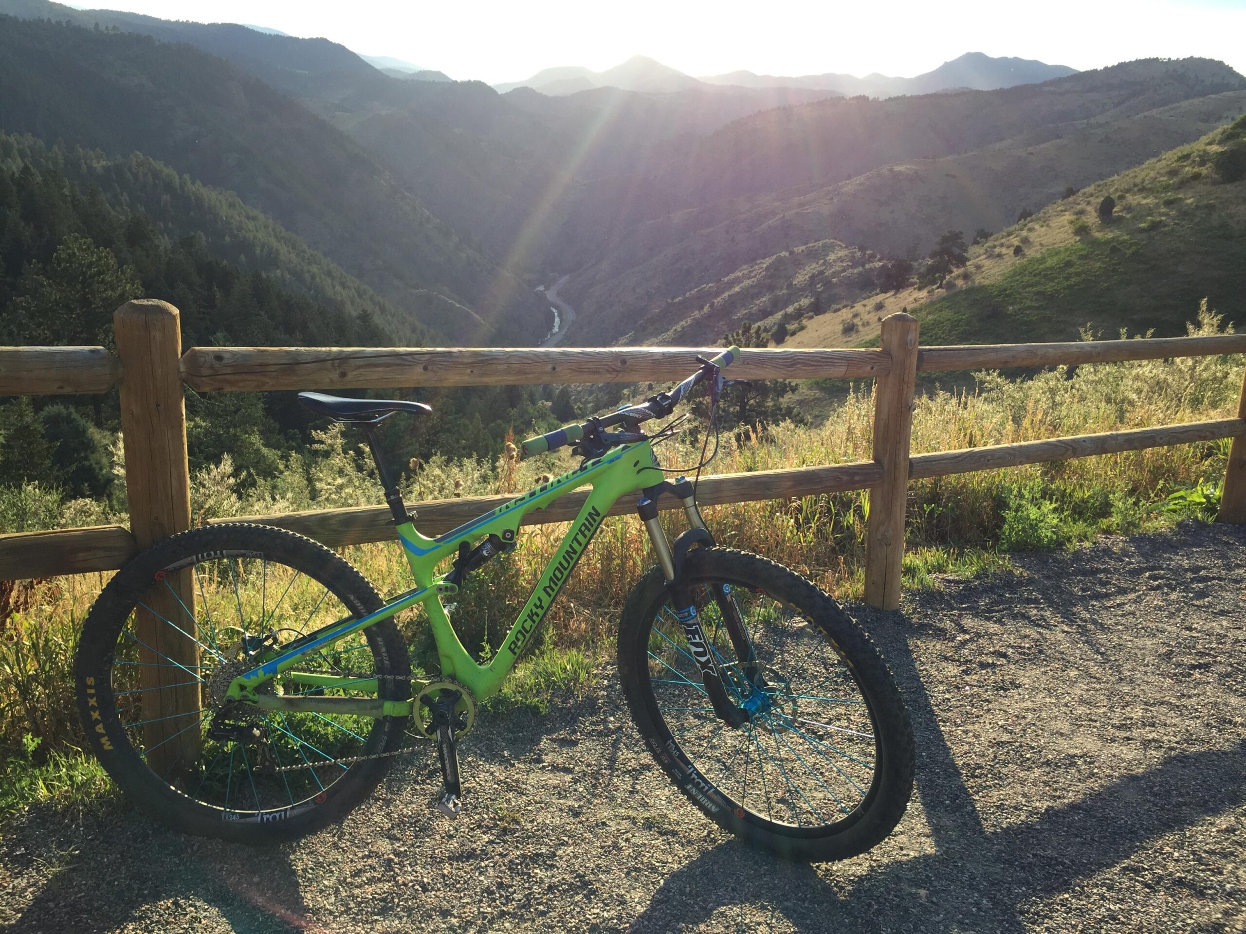 Mountain bike parked near a wooden fence overlooking a scenic valley and rolling hills. The sun is setting in the background, casting a warm glow over the landscape. Chimney Gulch mountain bike trail.