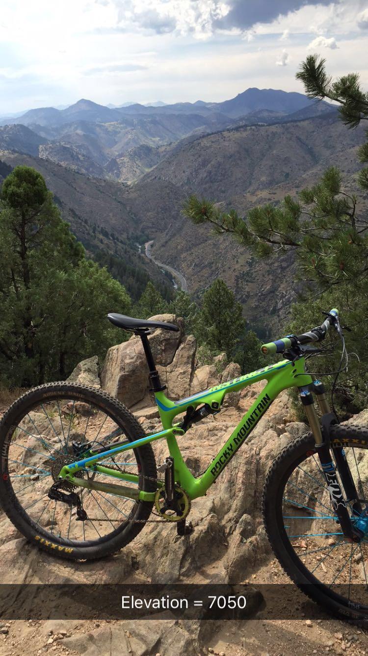 A green mountain bike is positioned on rocky terrain with a scenic view of mountains and valleys in the background. The elevation is noted as 7050 feet. Pines are visible in the foreground, and the sky is partly cloudy. Chimney Gulch mountain bike trail.