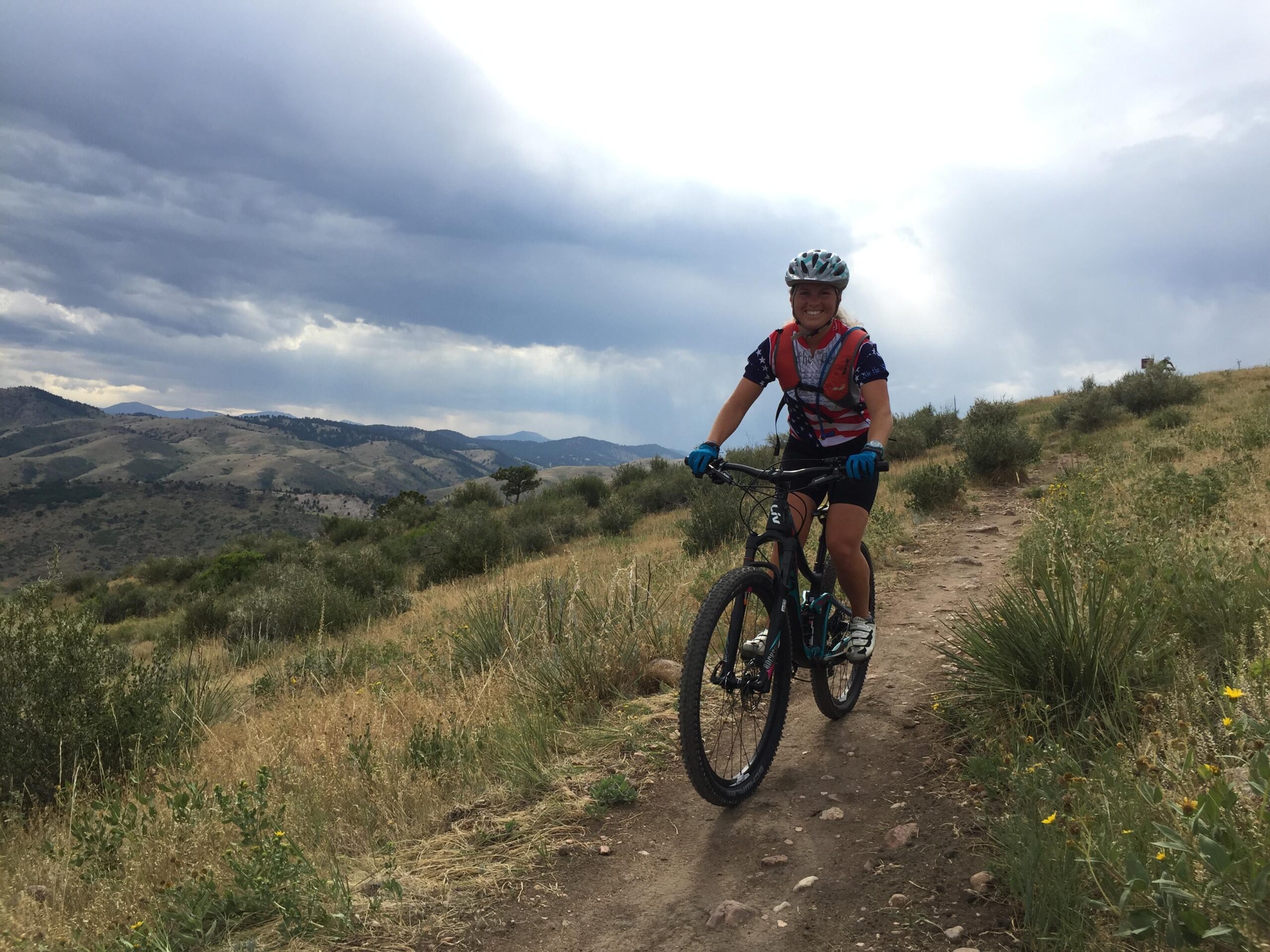 A person mountain biking on a dirt trail in a grassy landscape, with rolling hills and clouds in the background. The cyclist is wearing a helmet and a colorful jersey, smiling while riding their bike. Green Mountain mountain bike trail.