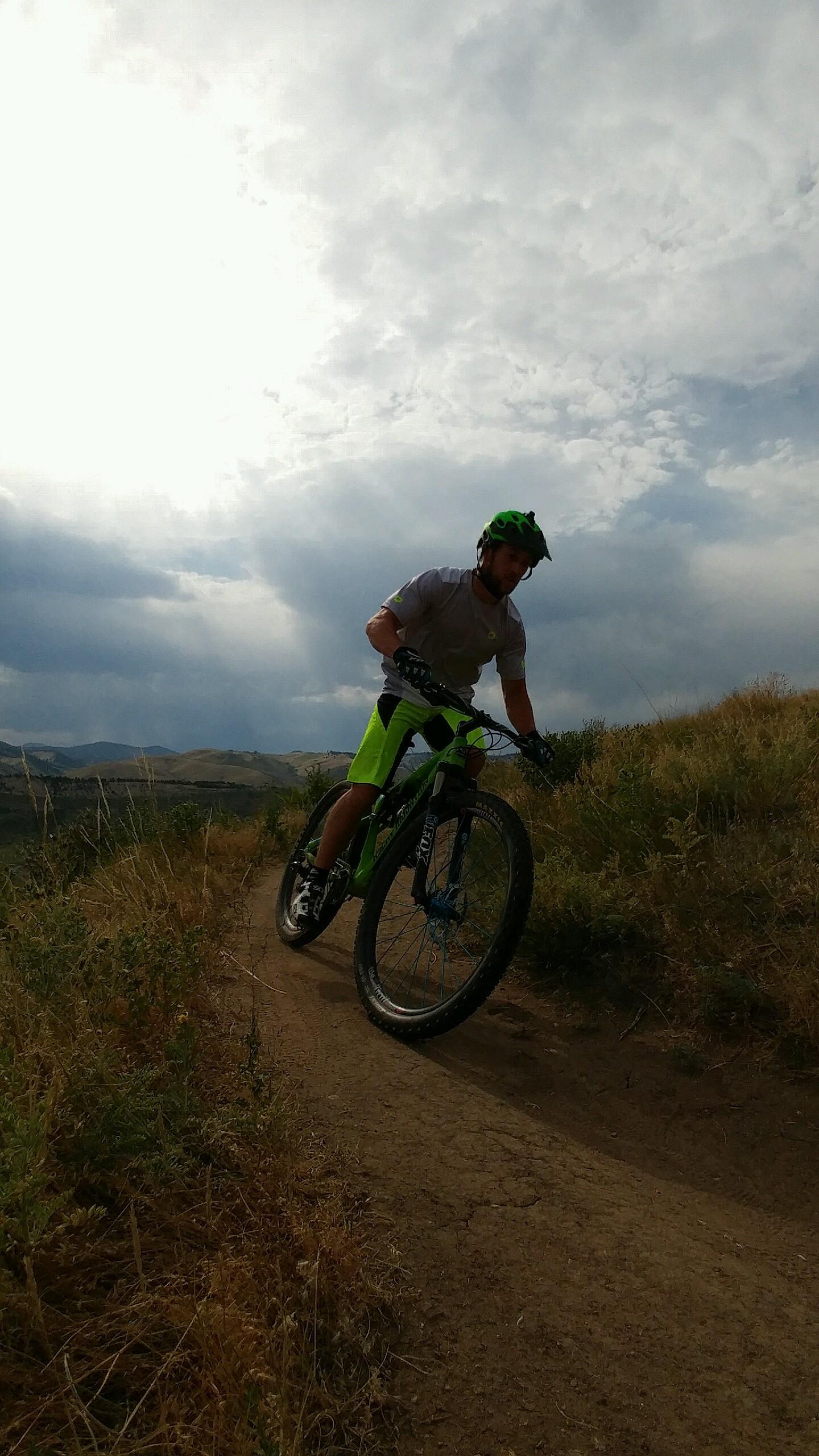 A mountain biker in a light gray shirt and bright green shorts rides along a dirt trail surrounded by tall grass and hills under a cloudy sky. Green Mountain mountain bike trail.