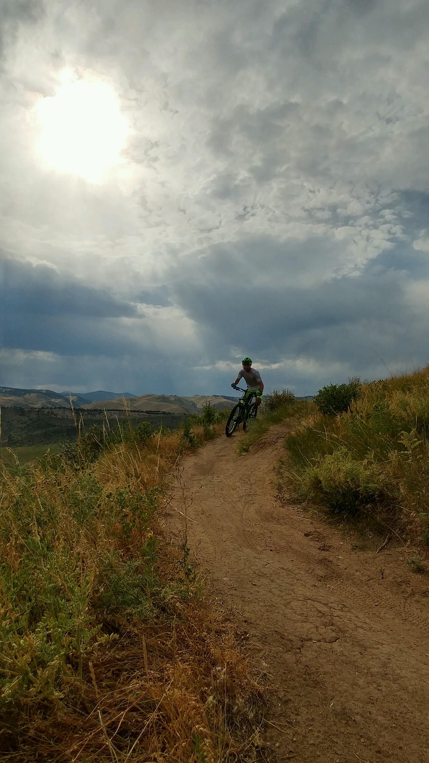 A mountain biker riding along a dirt trail surrounded by tall grass and shrubs, under a cloudy sky with sunlight breaking through. The landscape features rolling hills in the background. Green Mountain mountain bike trail.