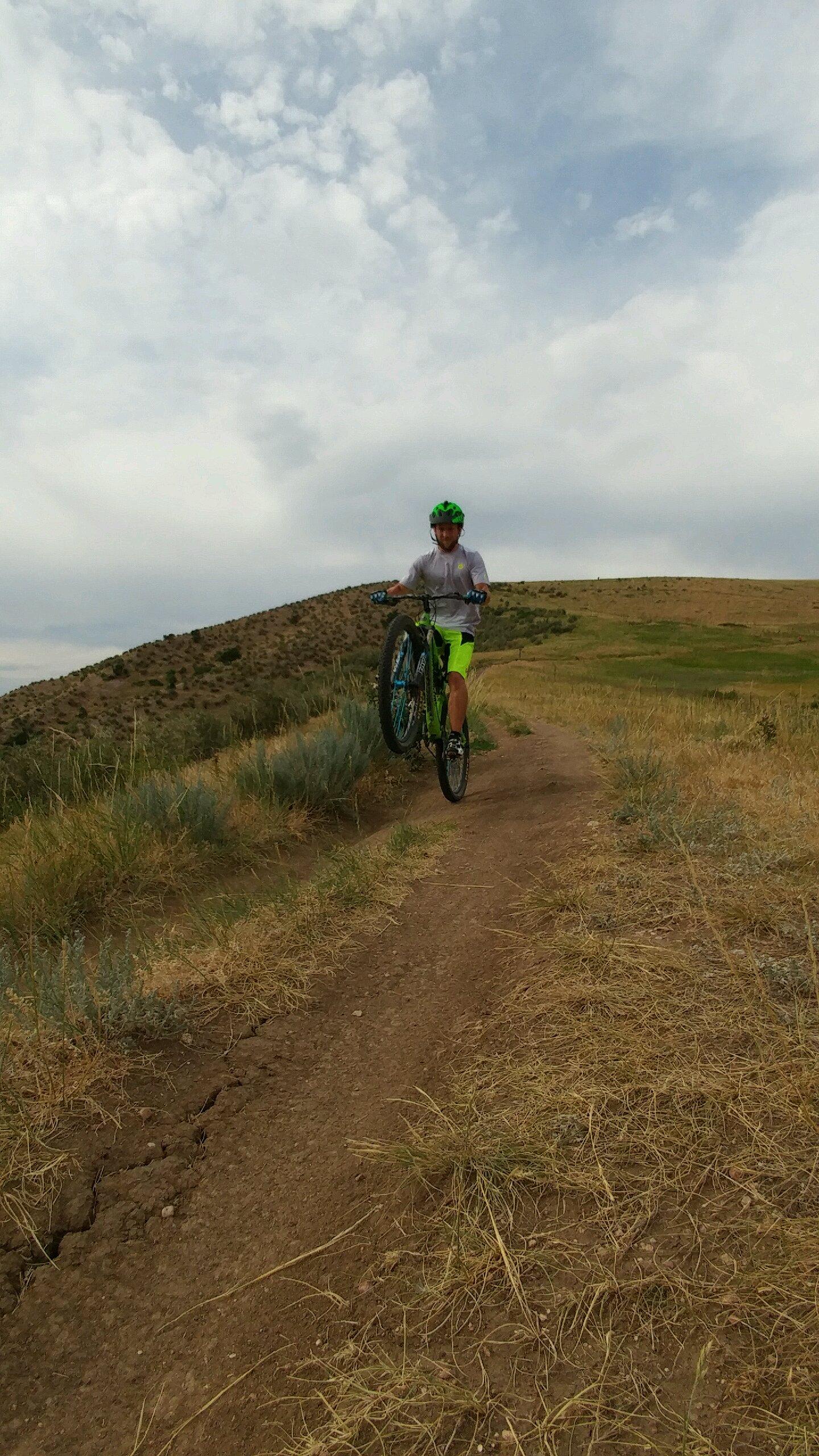 A person is riding a mountain bike on a dirt trail, performing a wheelie. The cyclist wears a bright green helmet and neon yellow shorts, surrounded by dry grass and shrubs, with rolling hills in the background under a partly cloudy sky. Green Mountain mountain bike trail.