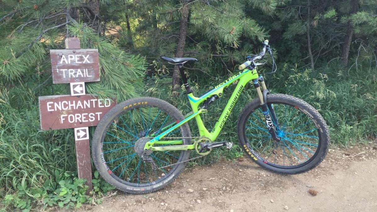A green mountain bike leaning against a wooden trail sign pointing to "Apex Trail" and "Enchanted Forest," surrounded by lush greenery and pine trees. Apex Park mountain bike trail.